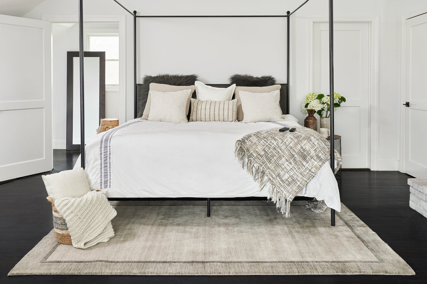 Black metal four-poster bed with white linens, beige pillows, and a cozy gray textured throw on a light beige rug.