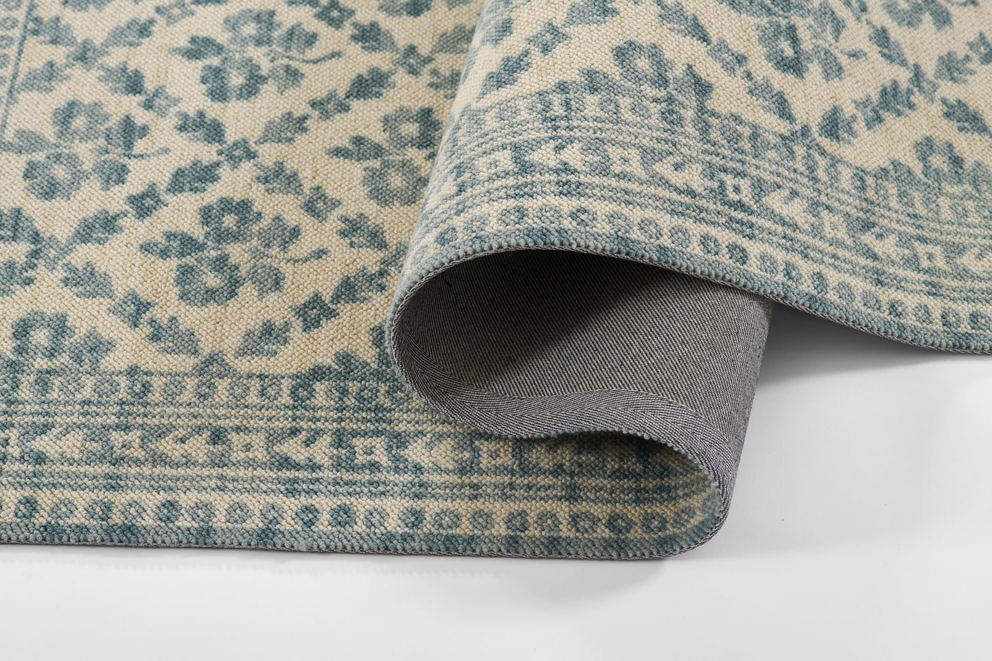 Beige and blue woven rug with a floral pattern, rolled edge reveals grey backing.