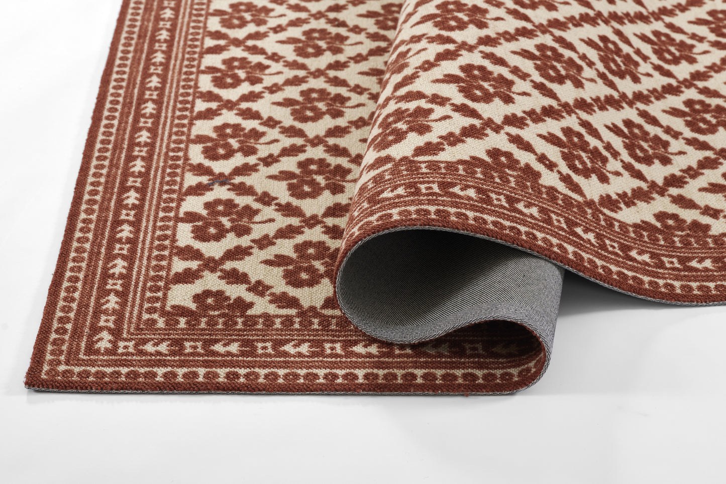 Red and cream ornate woven rug with floral motif and decorative border, rolled to show grey backing.