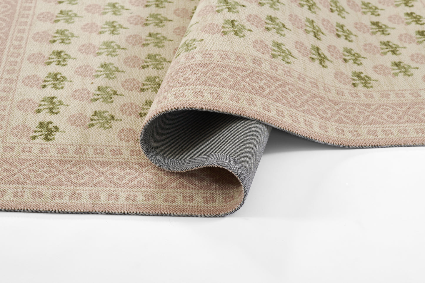 Pink patterned area rug with green floral motifs and geometric borders, rolled to reveal grey fabric backing.