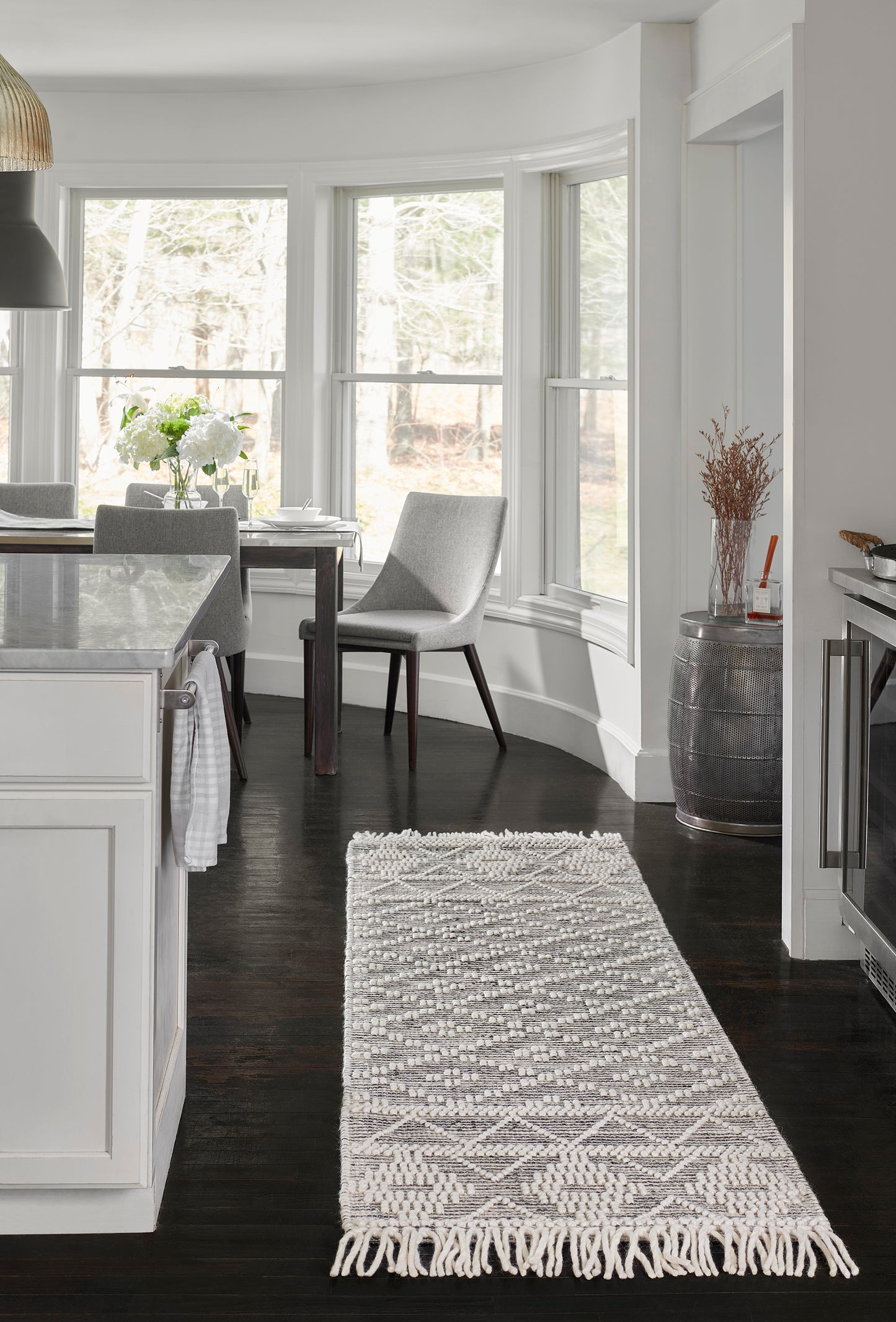 Bright white kitchen with curved bay windows, dark hardwood floors, a gray woven area rug with fringe, and a dining area.