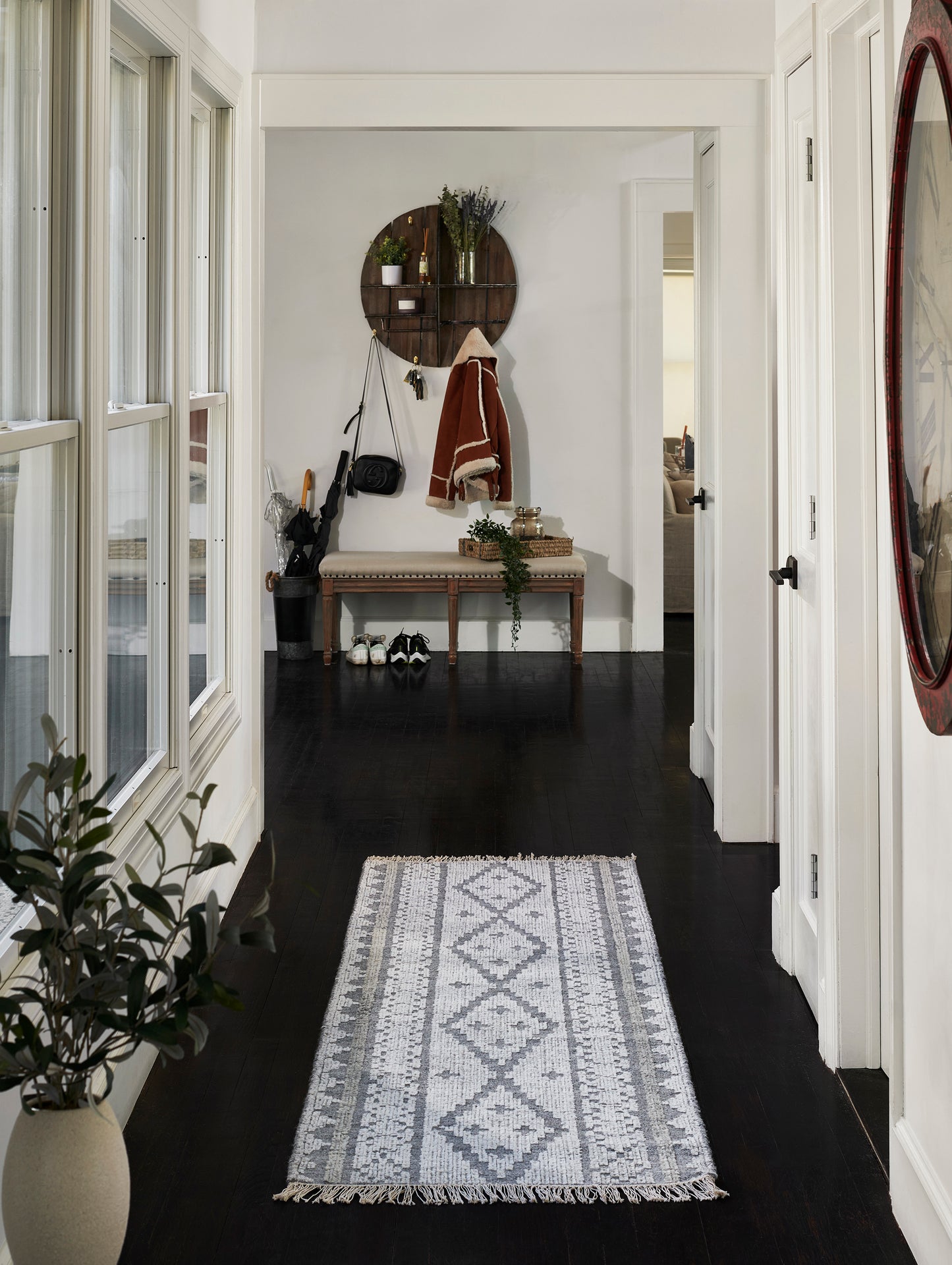 Gray geometric rug runner with fringe on dark hallway; white walls, bench, round wooden shelf, coats, plants, and a red circular mirror.