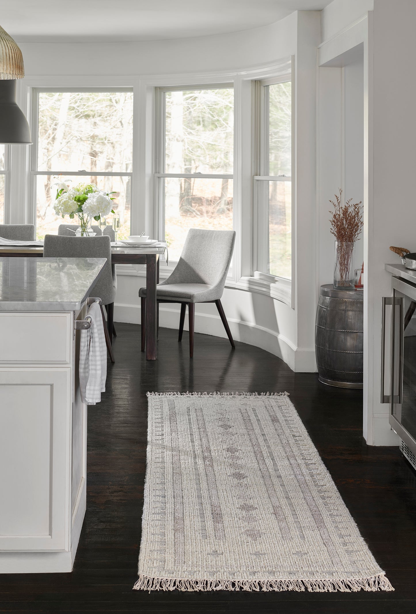 Ivory woven runner rug with fringe ends on dark wood floor beside white kitchen cabinets, bay window with grey chairs.