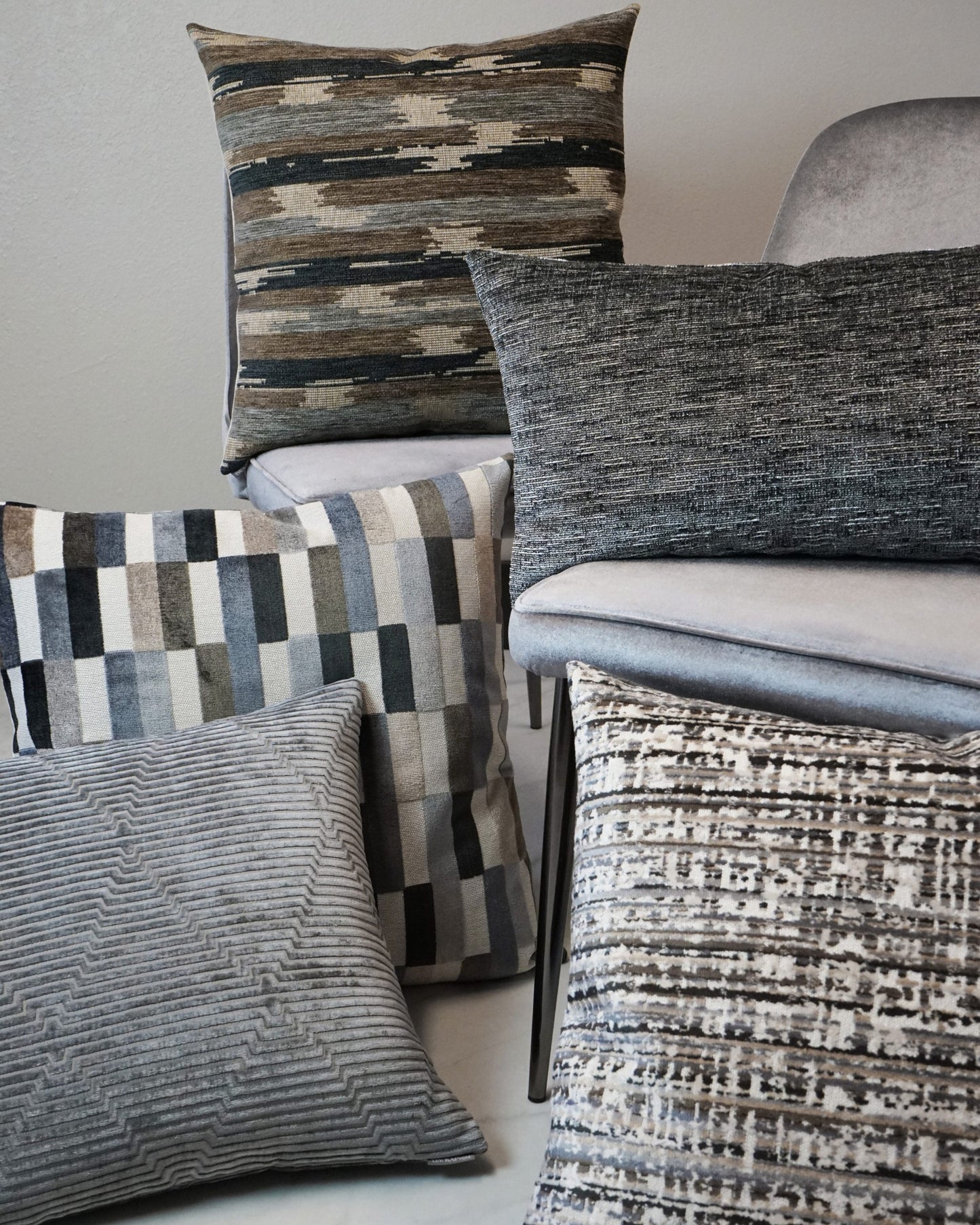 Decorative throw pillows in gray, taupe, and brown with geometric, striped, and woven textures, stacked near a gray chair.