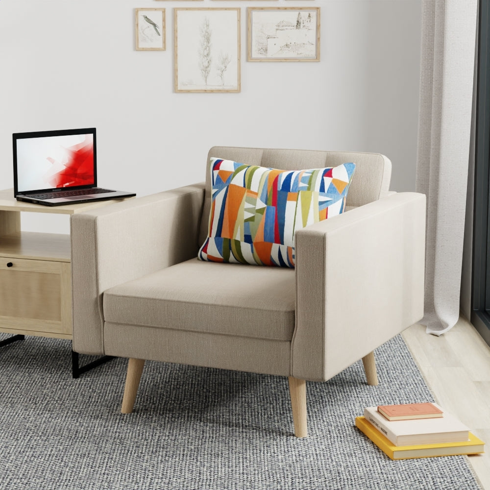 Beige upholstered armchair with light wood legs, colorful geometric cushion, in a modern living room with a laptop on a side table.