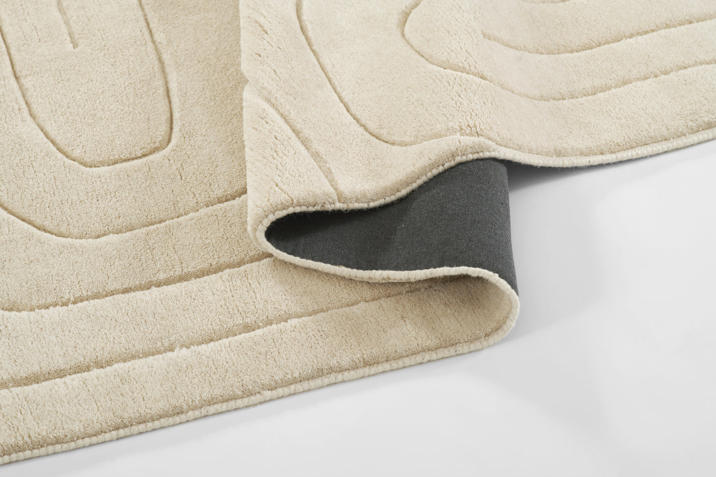 Beige soft-pile rug with abstract curved-line pattern; folded corner reveals dark gray underside.