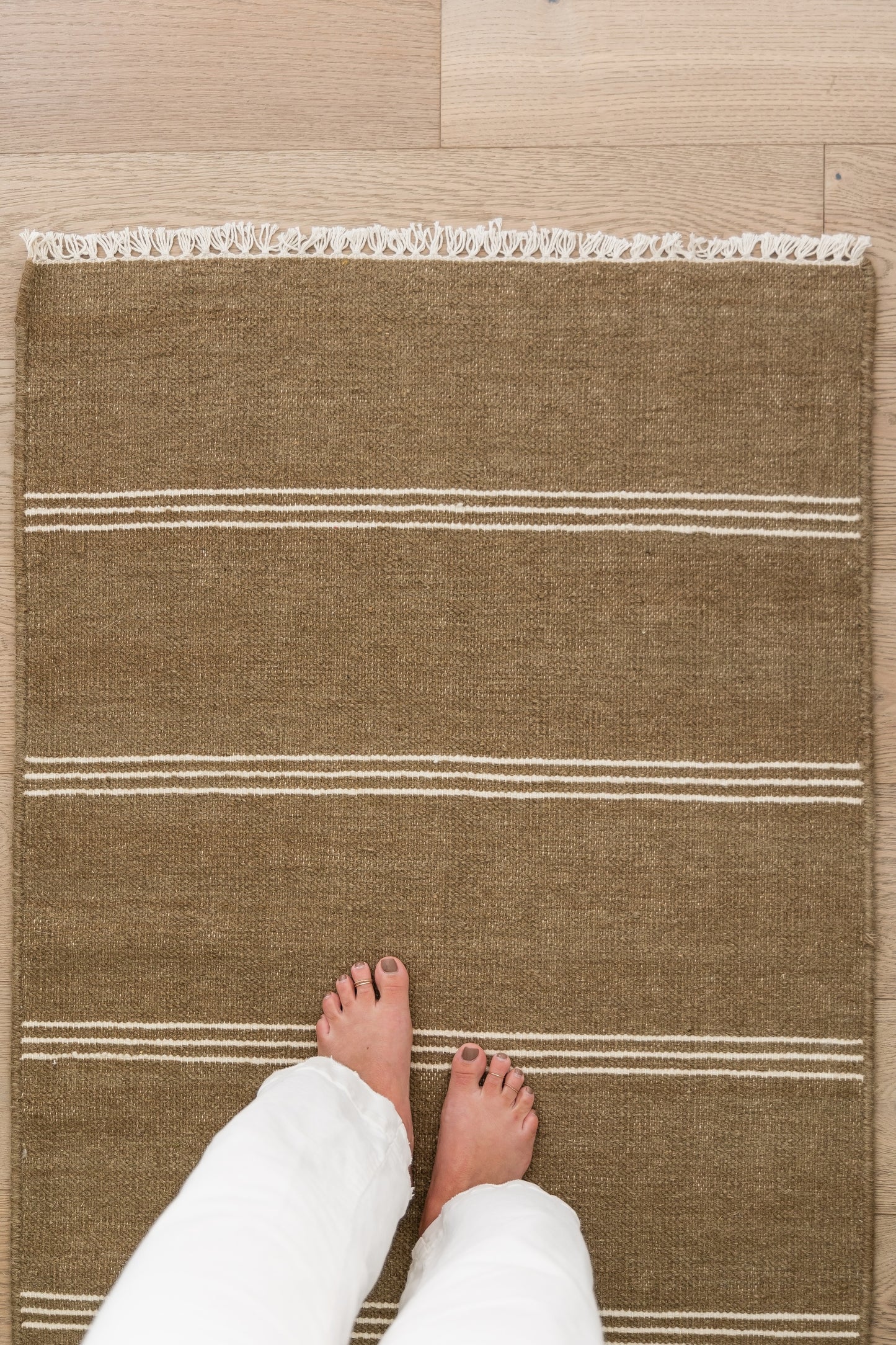 Brown jute rug with white stripes and fringe along top edge, rectangular on wood floor, feet for scale.