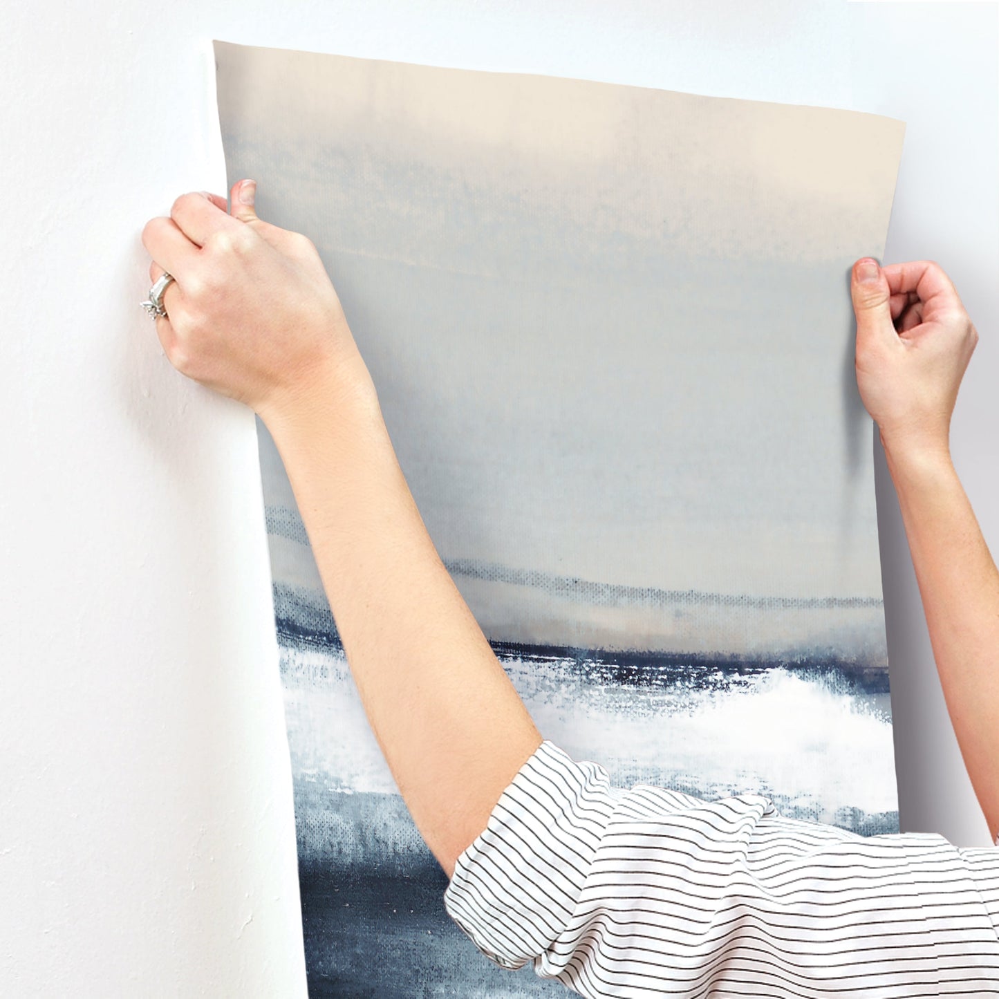 Hands in a striped shirt install a wall mural featuring an abstract blue-gray seascape with a beige horizon on a wall.