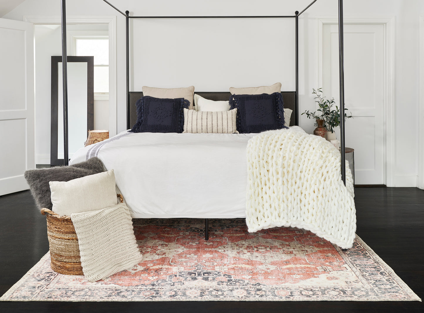 Black metal four-poster bed with white linens, navy & beige pillows, chunky knit white throw; woven basket & Persian rug.