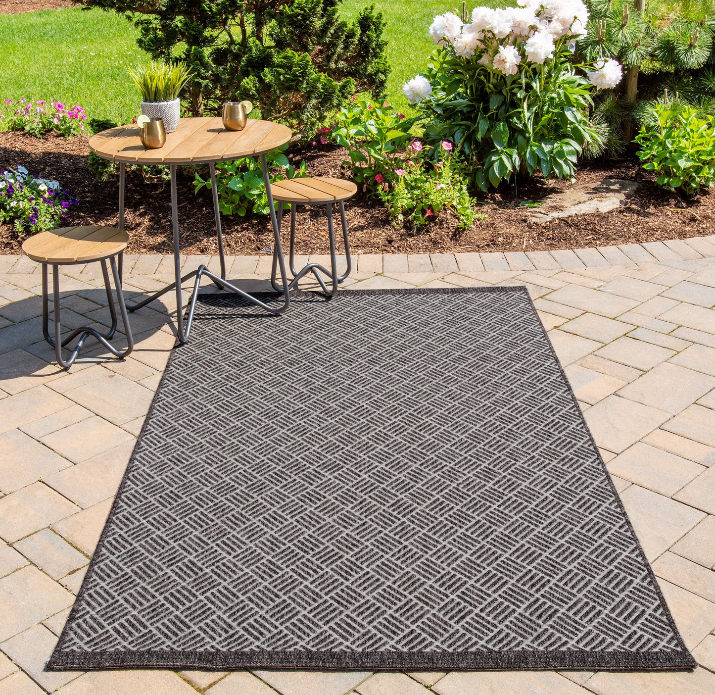 Grey outdoor runner rug (2' x 6') with a diamond pattern on a patio beside a round wooden table and stools, with a lush garden backdrop.