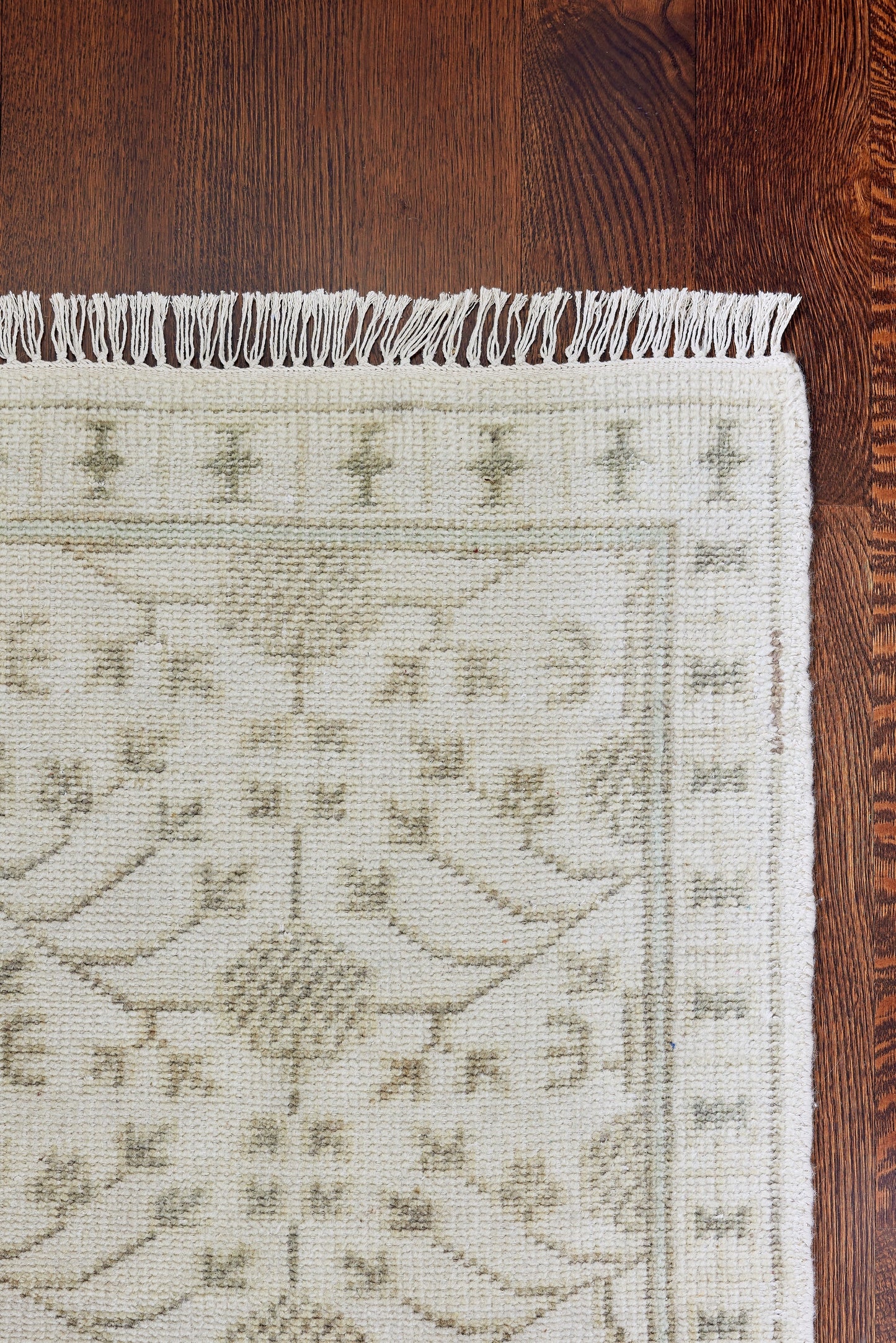 Cream woven rug with pale green/gray geometric floral motif, fringed border along top, on dark wood floor.