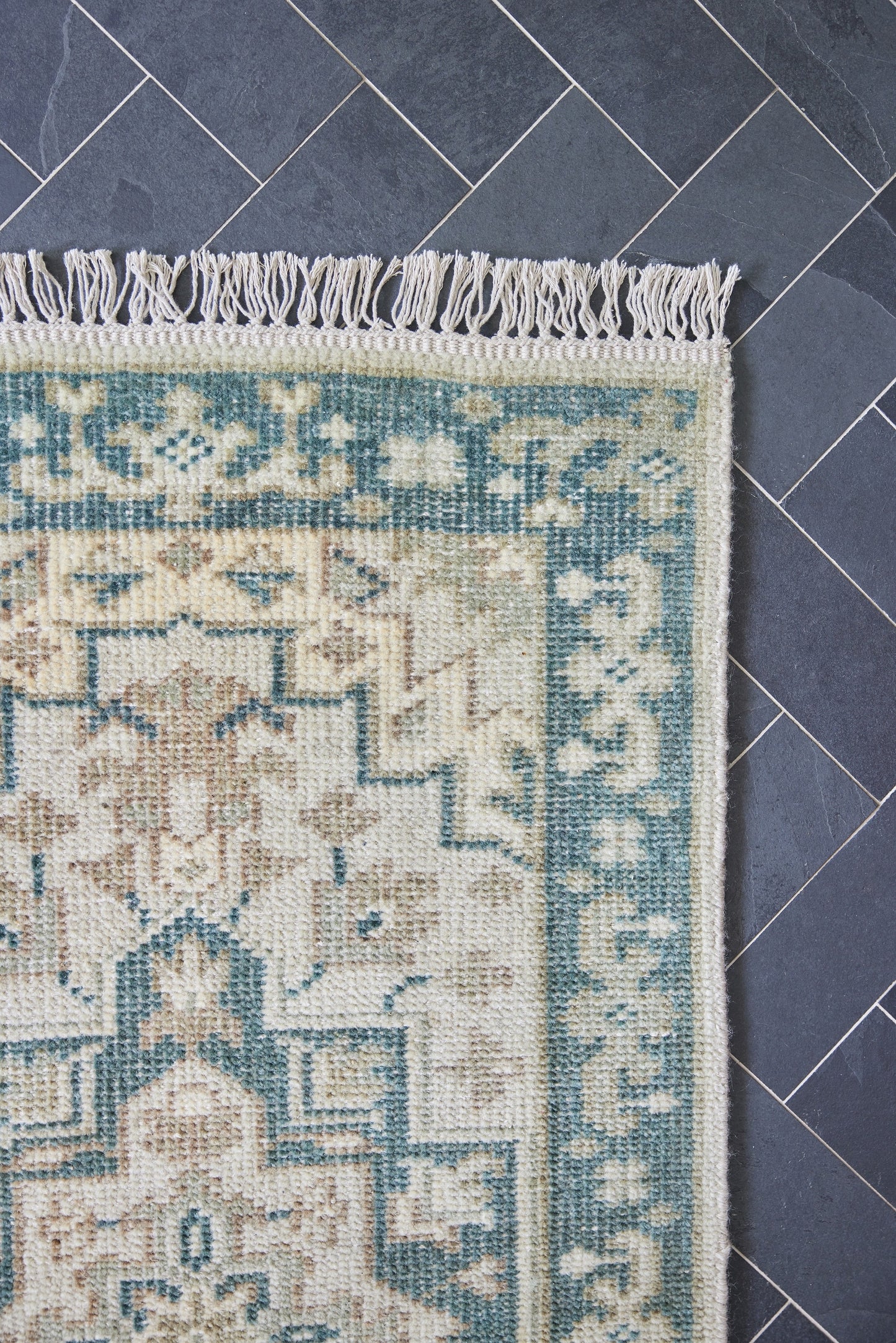 Corner of a cream and teal geometric rug with fringe on a dark slate tile floor.