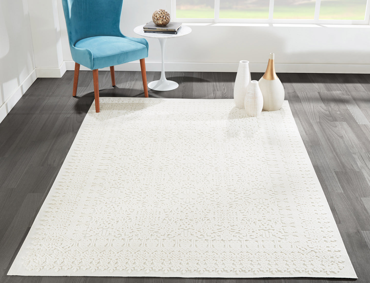 Ivory textured area rug with a subtle raised pattern on a dark wood floor; blue chair and white side table with decorative vases.