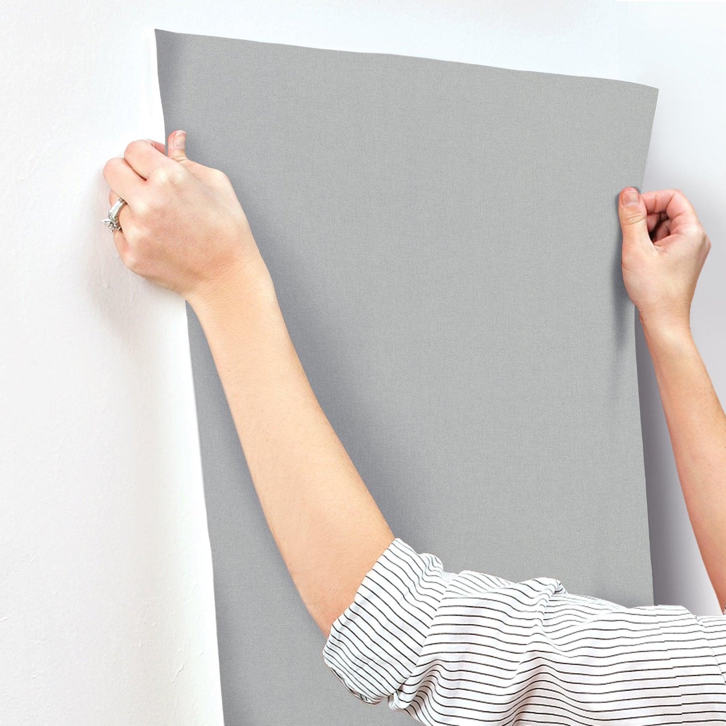 Hands in a striped shirt pull a large gray wallpaper panel against a white wall.