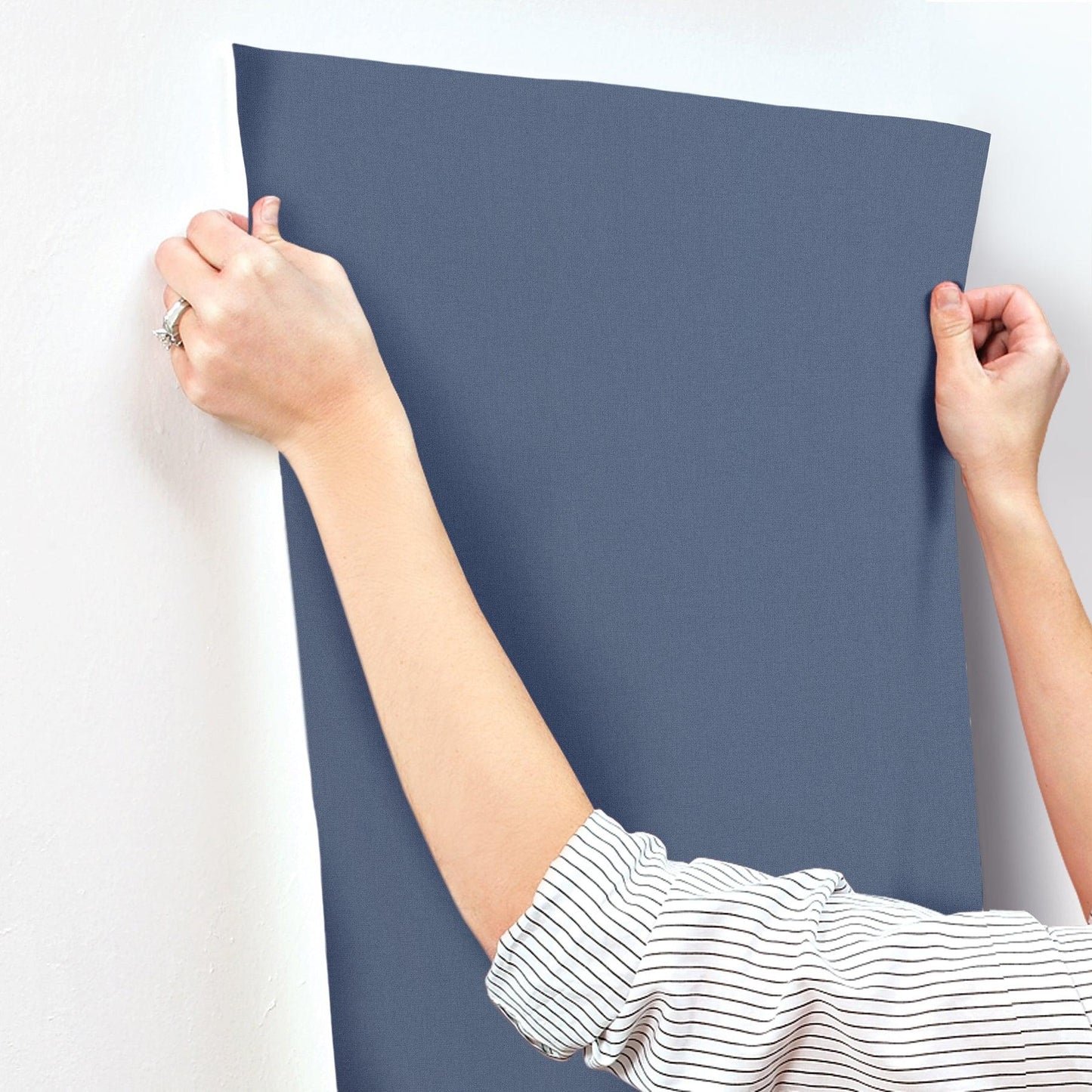 Rectangular slate-blue wallpaper being stretched onto a wall by hands; striped sleeves visible.