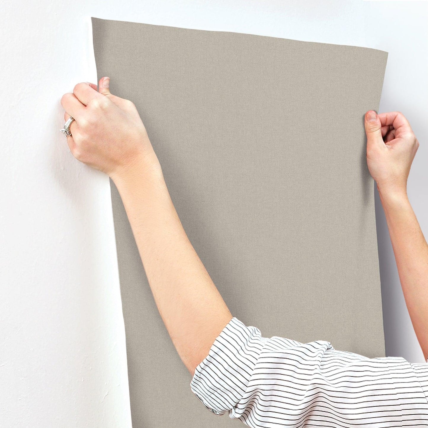 Beige wallpaper panel being hung on a wall; hands grip the top corners, striped sleeve visible.