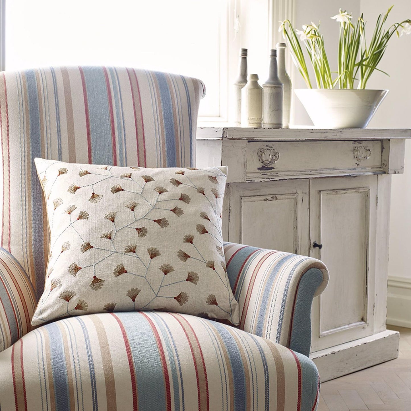 Pastel striped armchair with beige gingko-leaf cushion; rustic whitewash cabinet and potted lilies.