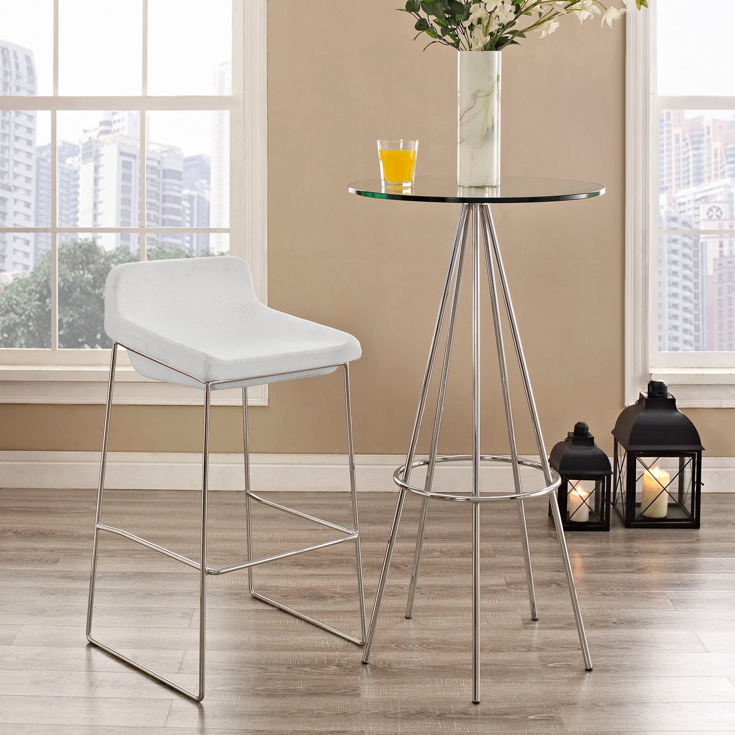 White cushioned bar stool with chrome legs, shown beside a round glass tripod table with a vase of white flowers and an orange drink.