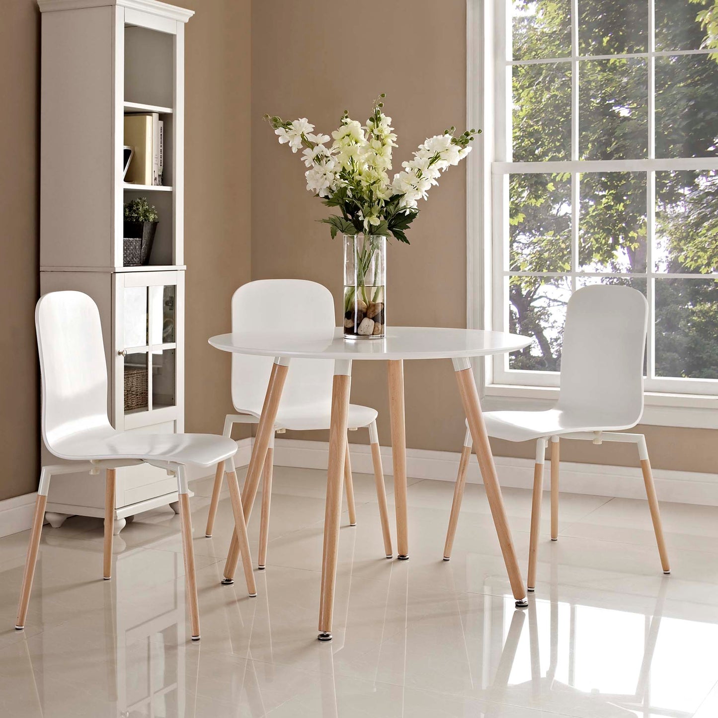 White round dining table with light wood legs, three white chairs, a vase of white flowers, in a sunlit dining room.