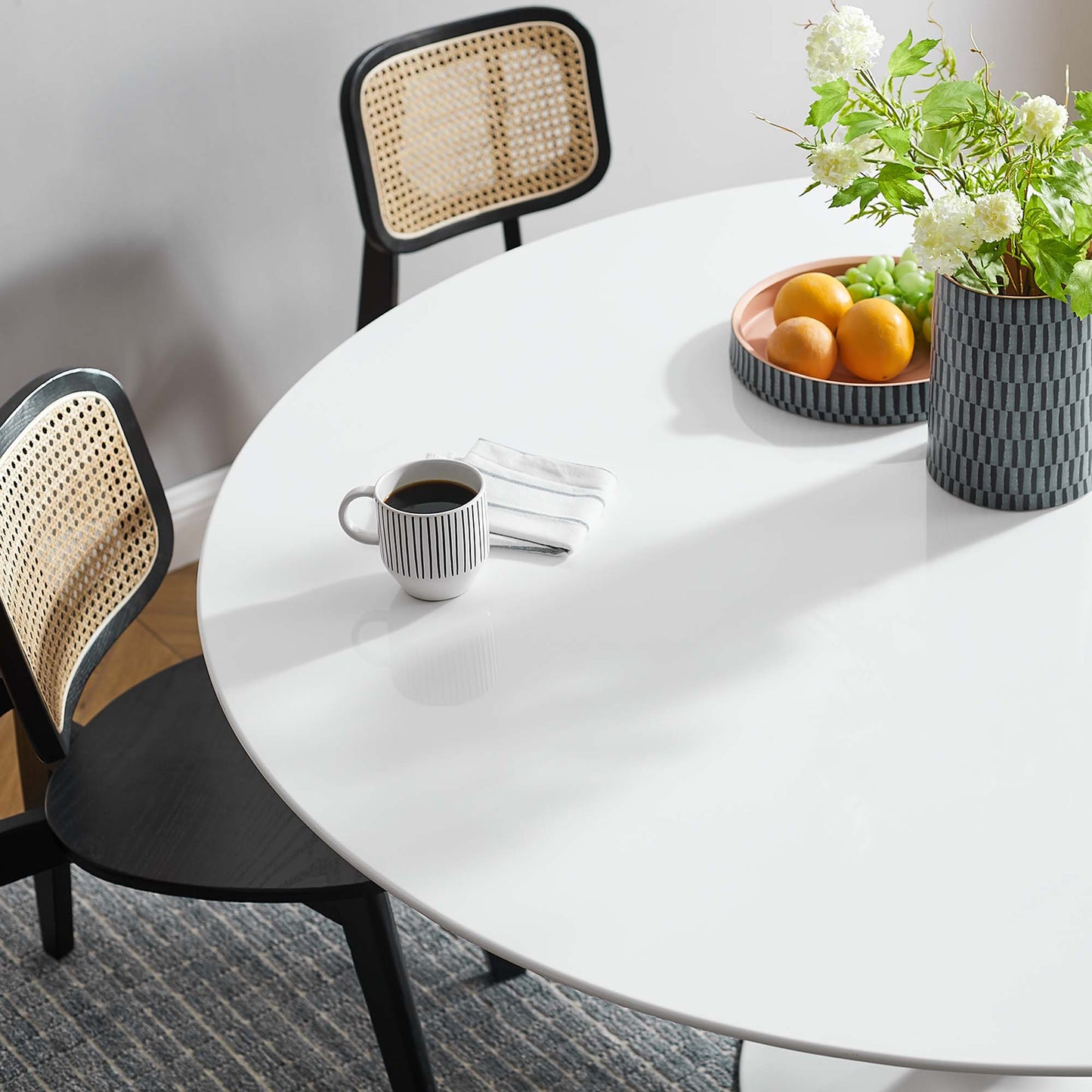 Round white table with black legs, caned-back chairs, striped mug, napkin, bowl of oranges, and a vase of white flowers.