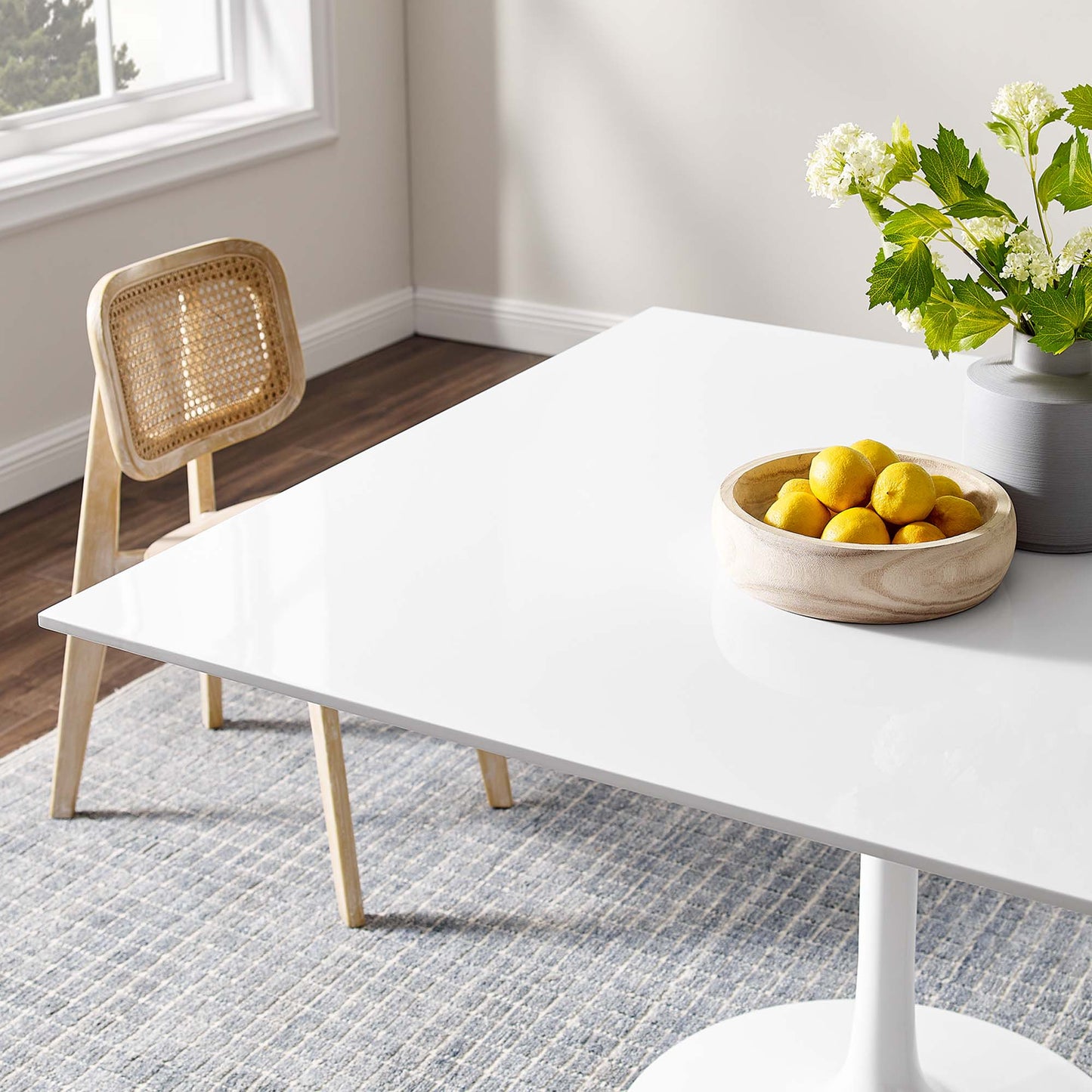 Glossy white rectangular dining table with wooden legs in a bright room; wicker chair, bowl of lemons, and a vase of white flowers on a grey rug.