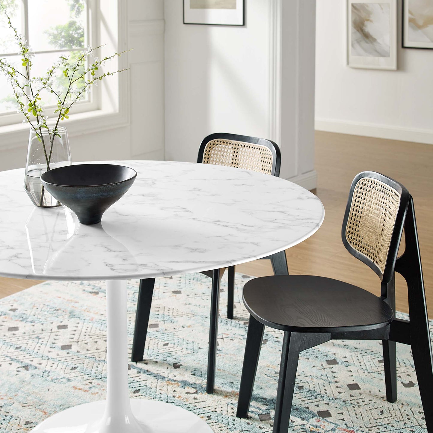 Round marble-top dining table with white pedestal base, black chairs with caned backs; modern dining set with vase and bowl.