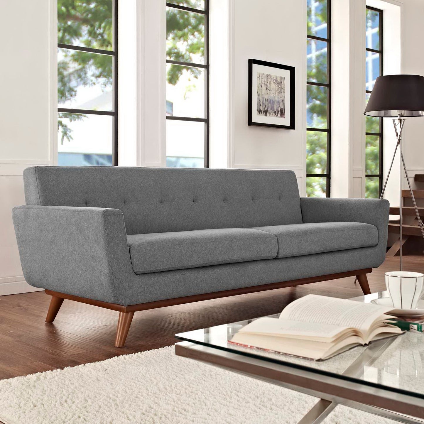 Grey mid-century sofa with tufted back, two cushions and angled wooden legs in a bright living room with large windows.