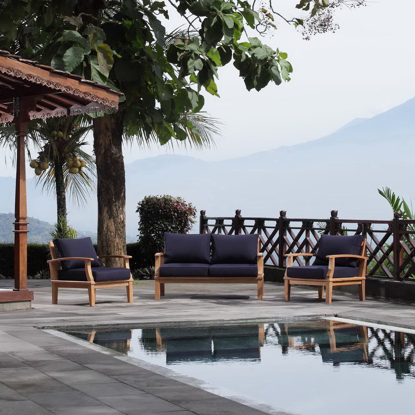 Three-piece outdoor lounge set in natural wood with navy cushions, poolside under palm trees with mountain view.