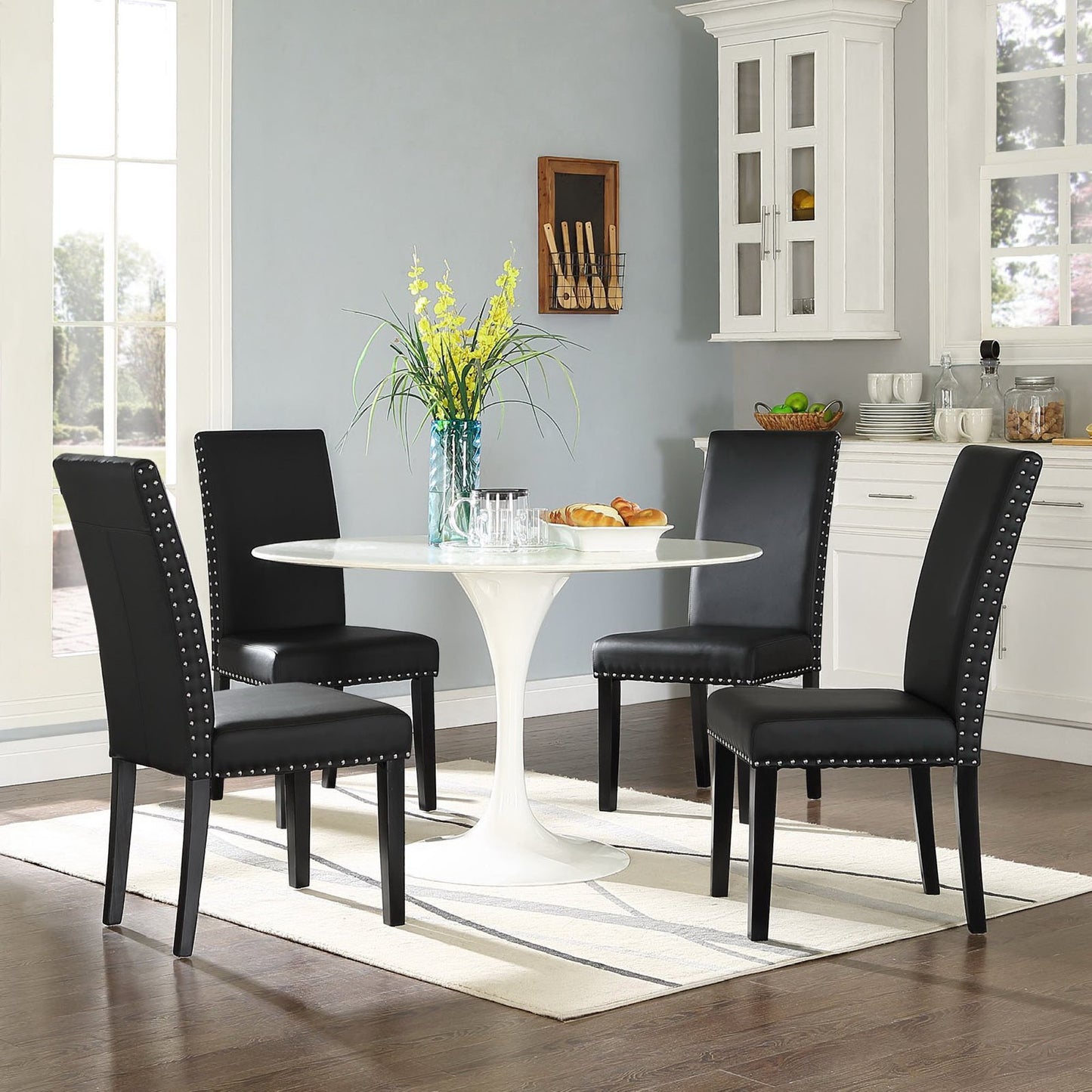 Round white tulip pedestal dining table with four black upholstered chairs with nailhead trim in a bright kitchen nook.