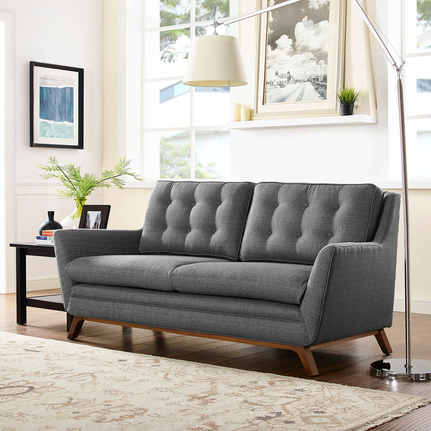 Gray mid-century tufted fabric loveseat with wooden legs in a bright living room with large windows and framed art.