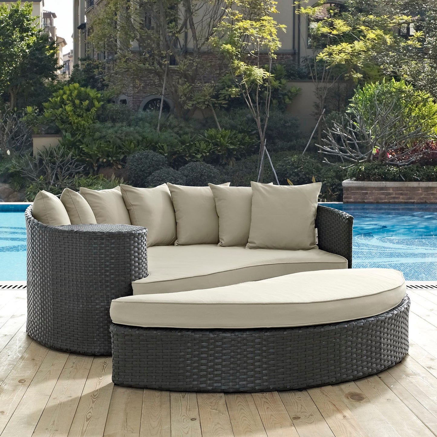 Round dark wicker outdoor daybed with light beige cushions, modular curved sections and backrest pillows by pool.