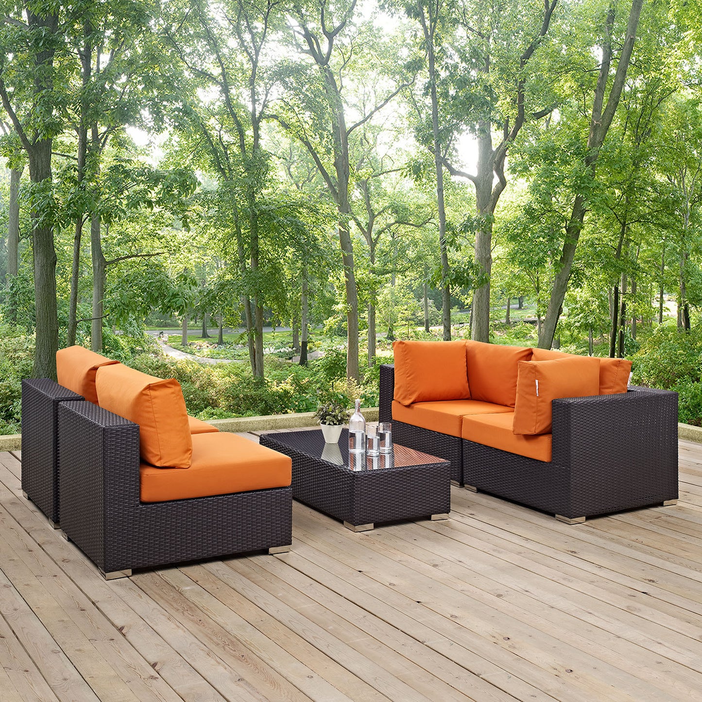 Espresso wicker outdoor sectional set with orange cushions and a glass coffee table on a wooden deck amid lush trees.