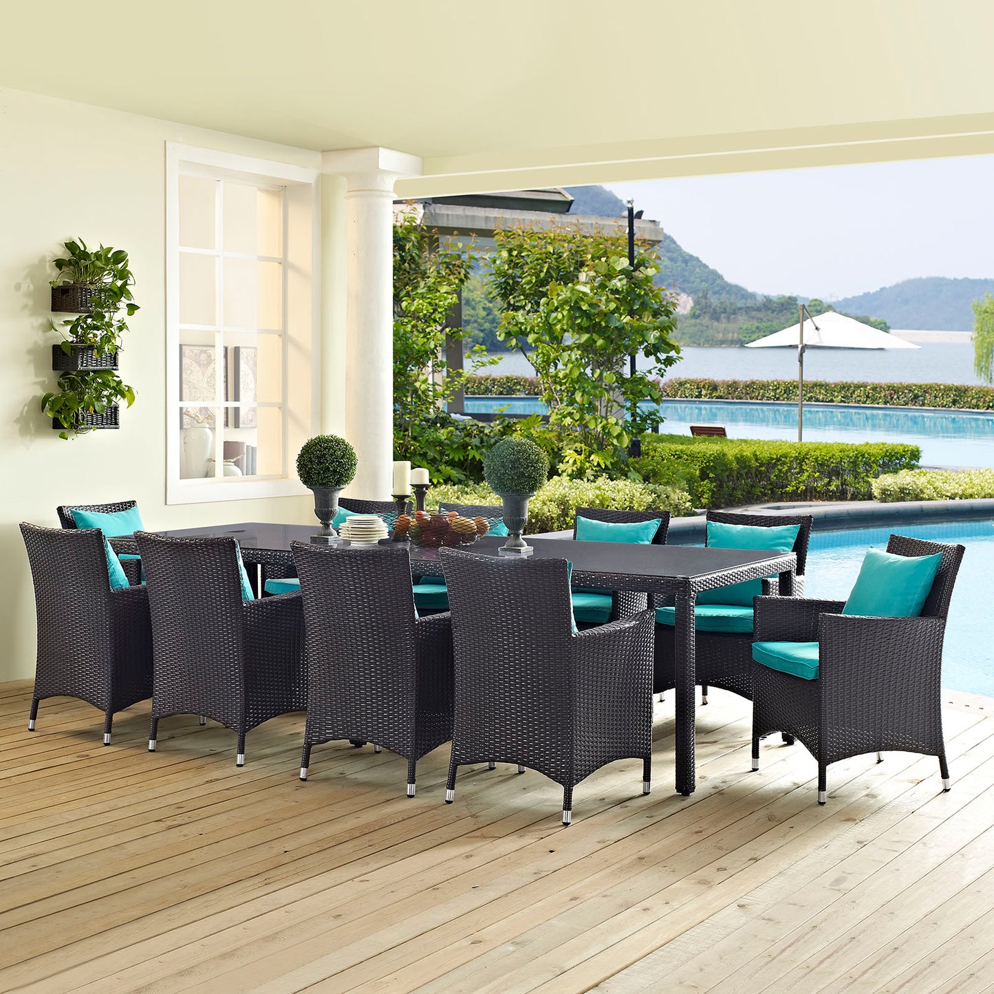 Rectangular dark brown wicker outdoor dining table with ten turquoise cushions on a wooden deck by a pool.