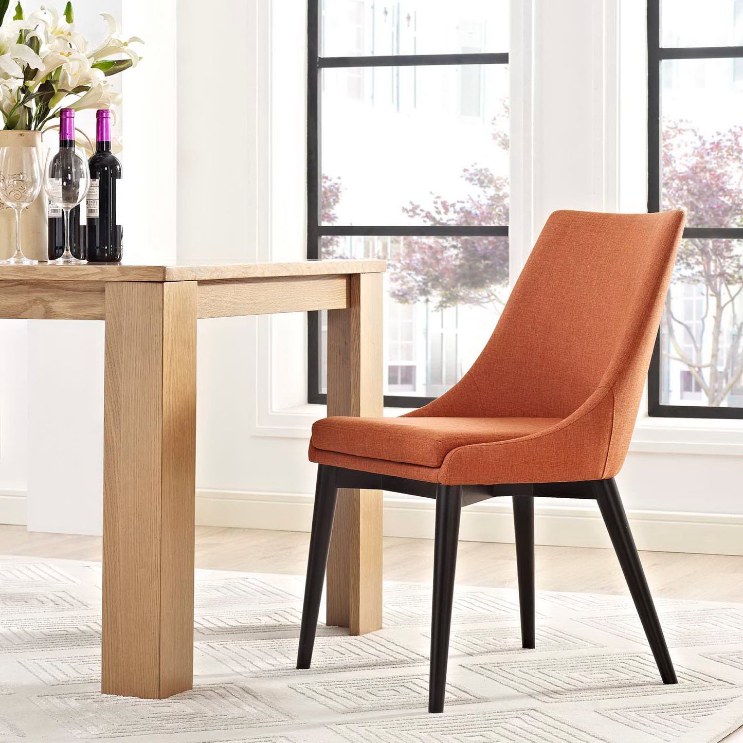 Orange fabric mid-century dining chair with black tapered legs beside a natural wood table in a bright dining room.