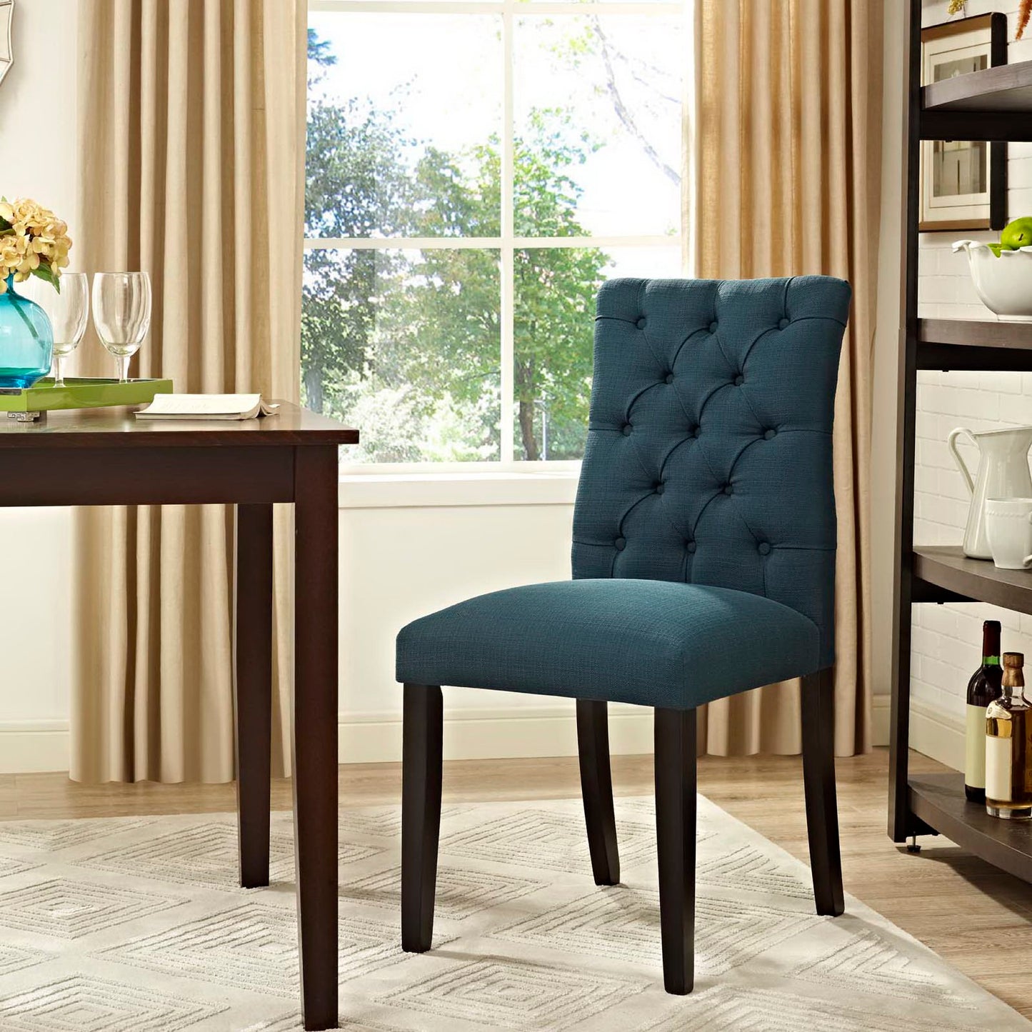 Blue button-tufted fabric dining chair with dark wood legs in a sunlit dining nook beside a wooden table and open shelving.