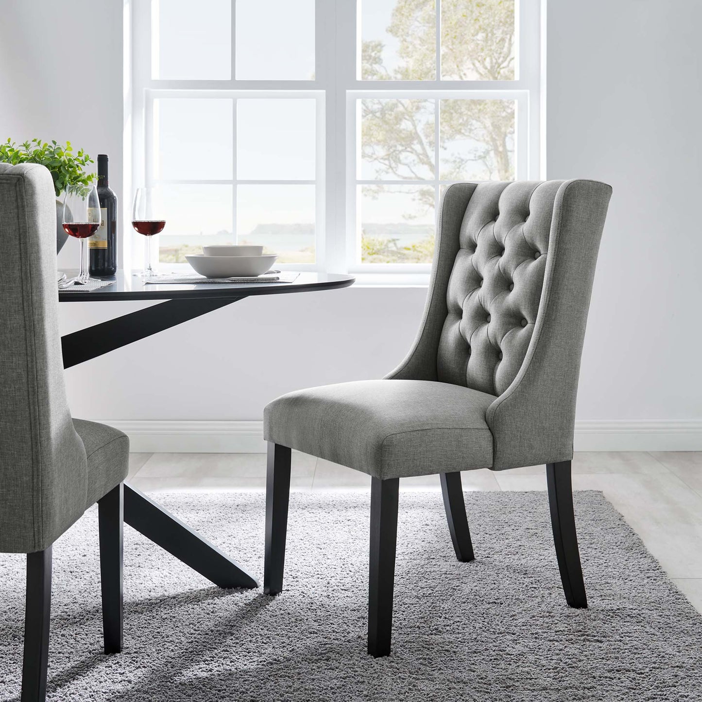 Pair of gray tufted dining chairs with dark wood legs beside a round black table in a bright windowed dining room.