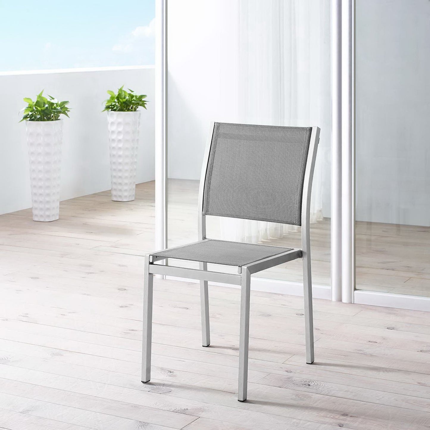 Silver-gray aluminum outdoor side chair with mesh seat and back on a light wooden deck, with glass doors and tall white planters.