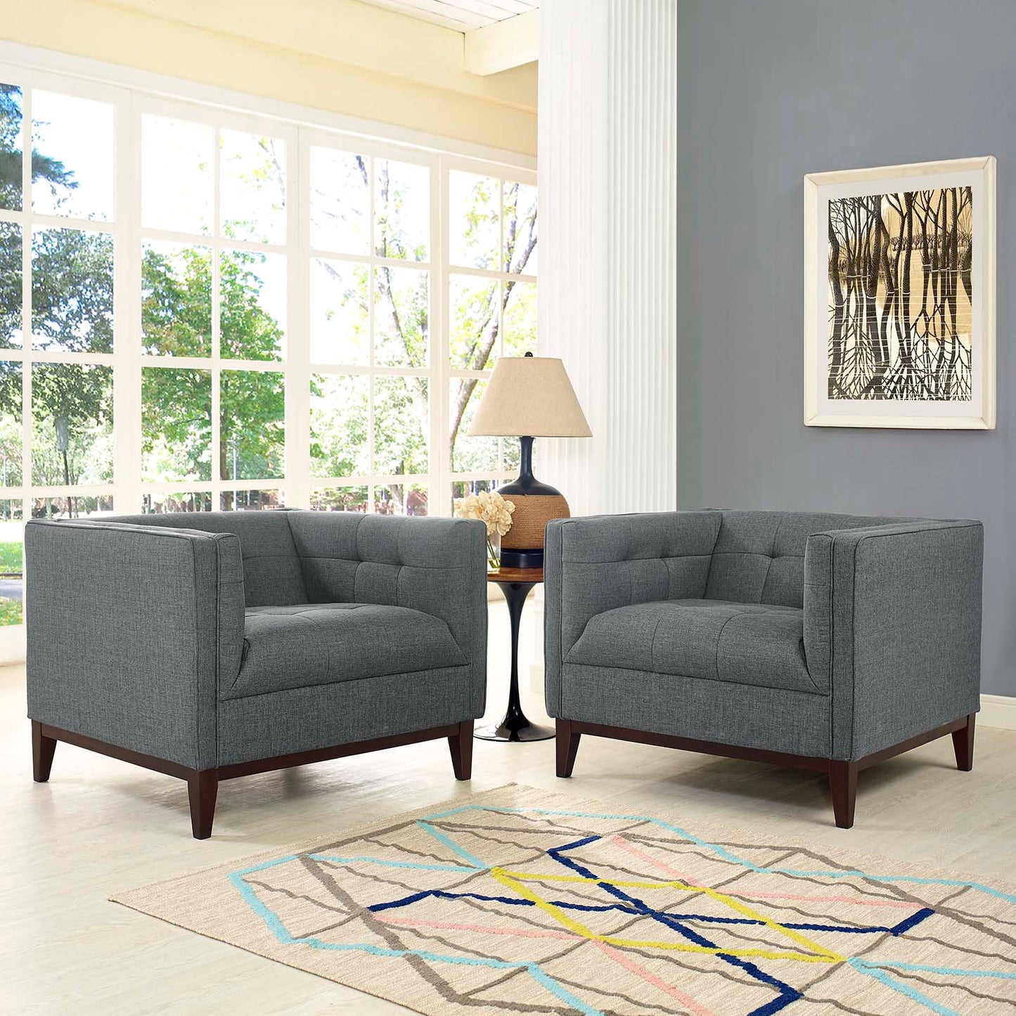 Two gray fabric armchairs with dark wood legs in a bright living room, between a round table and lamp; geometric rug, art.