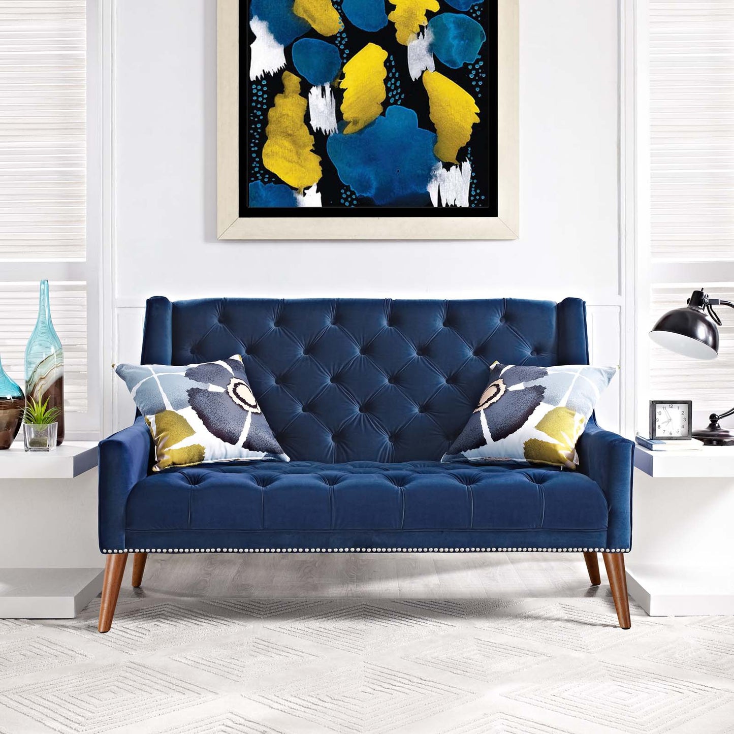 Navy tufted loveseat with nailhead trim and wooden legs, two geometric pillows; abstract blue-yellow art on white wall.