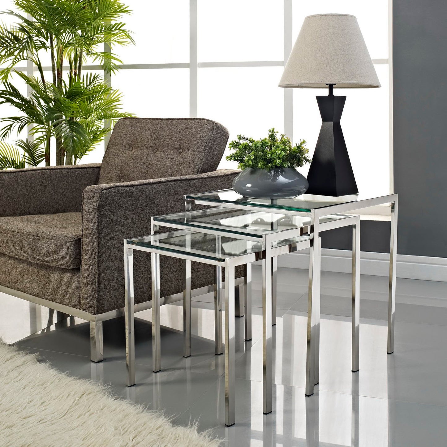 Chrome & glass nesting tables (set of 3) beside a brown upholstered sofa, with a lamp and greenery in a modern living room.