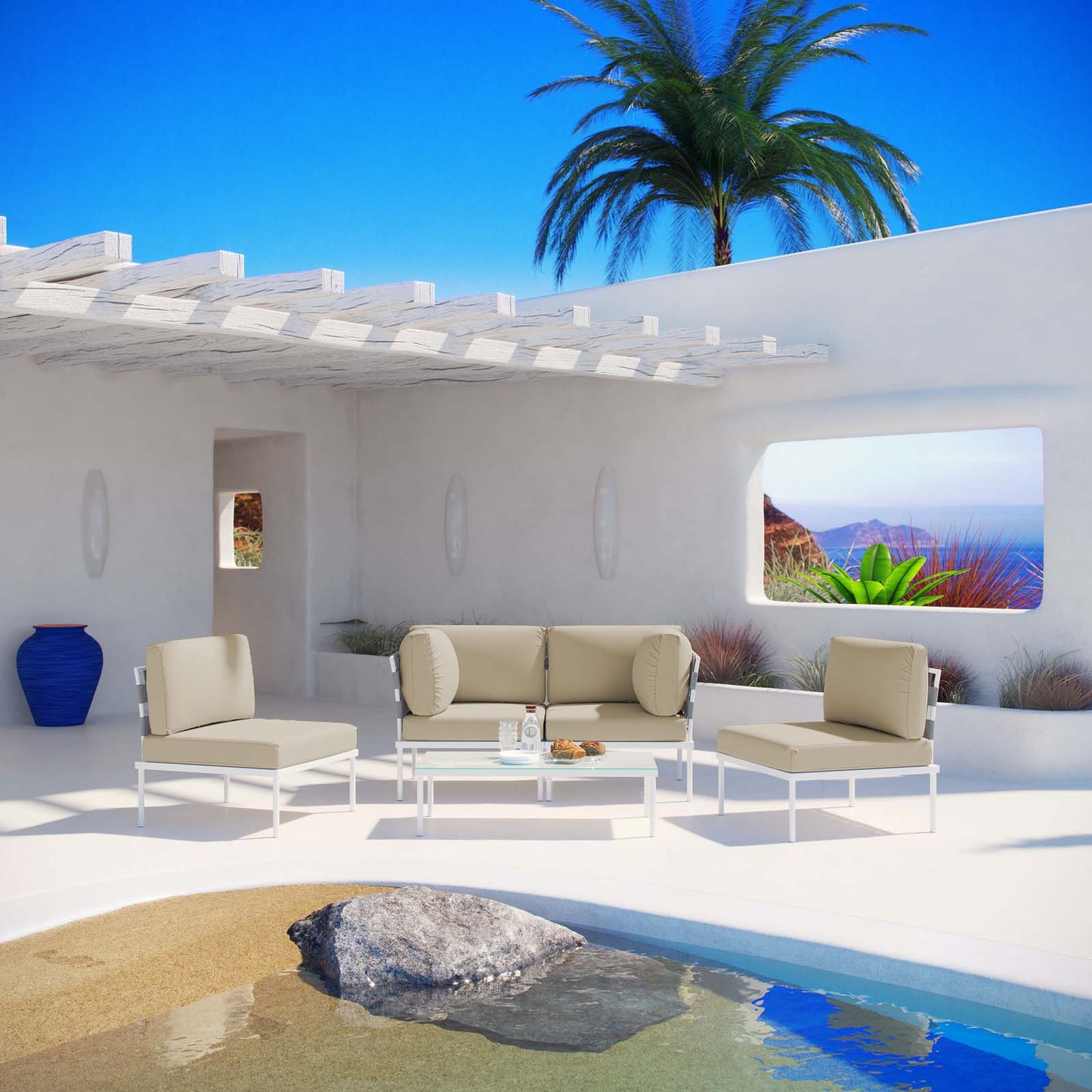 White outdoor seating set with beige cushions & bolster pillows on a sunlit stucco patio by a pool, palm tree & blue sky.