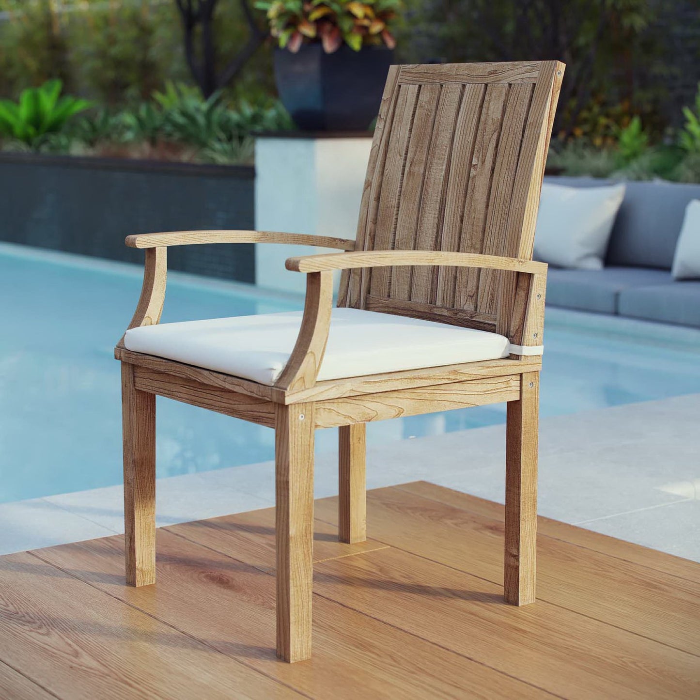 Natural finish wooden outdoor chair with white cushion, slatted back, curved armrests by poolside.