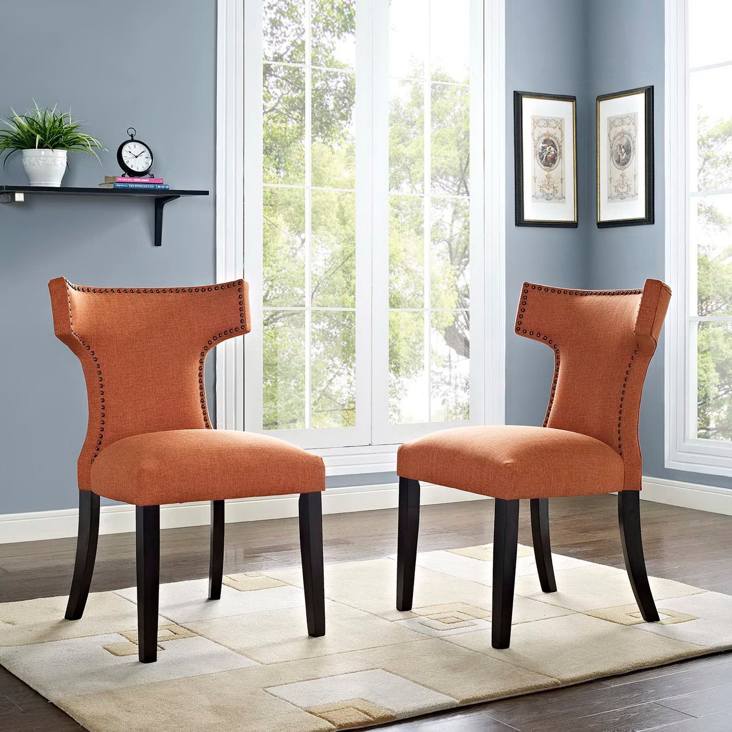 Pair of orange upholstered dining side chairs with nailhead trim, curved backs, and dark wood legs in a bright room with large windows.