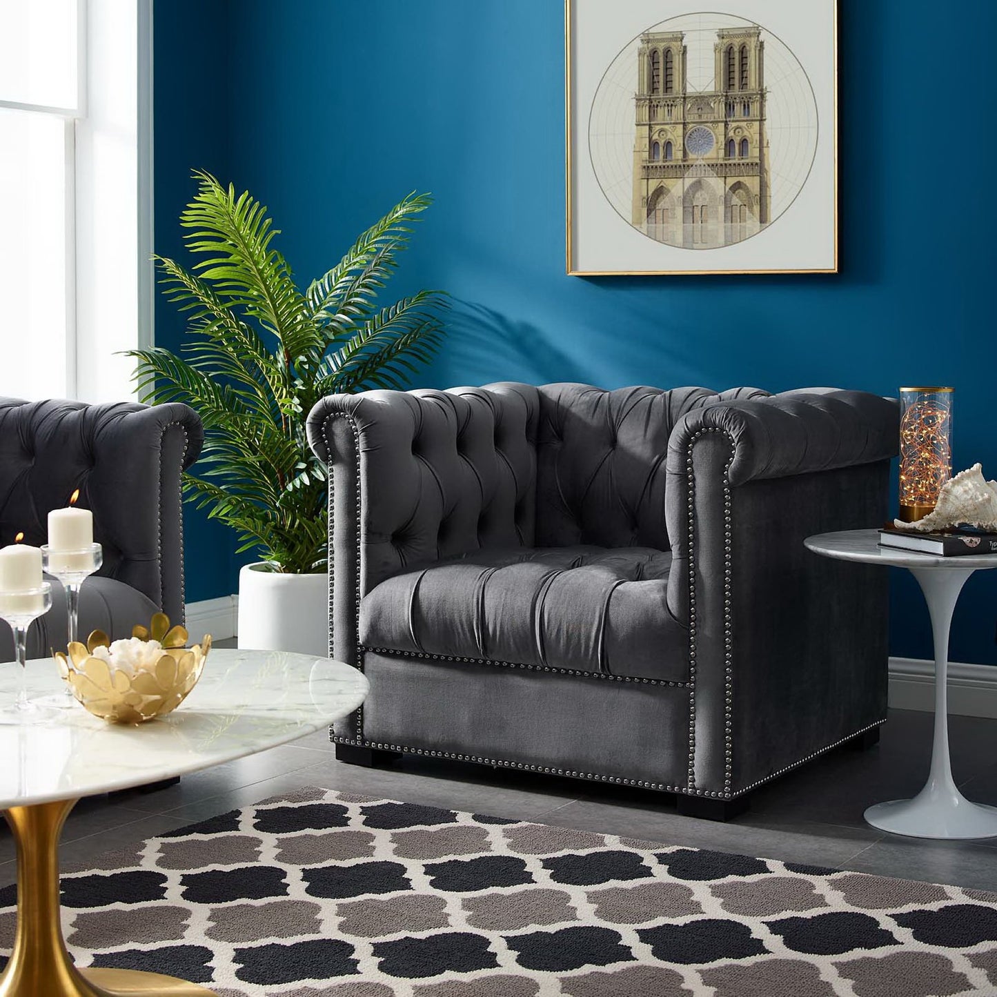 Dark gray tufted Chesterfield armchair with nailhead trim, blue wall, round marble coffee table, plant, geometric rug.