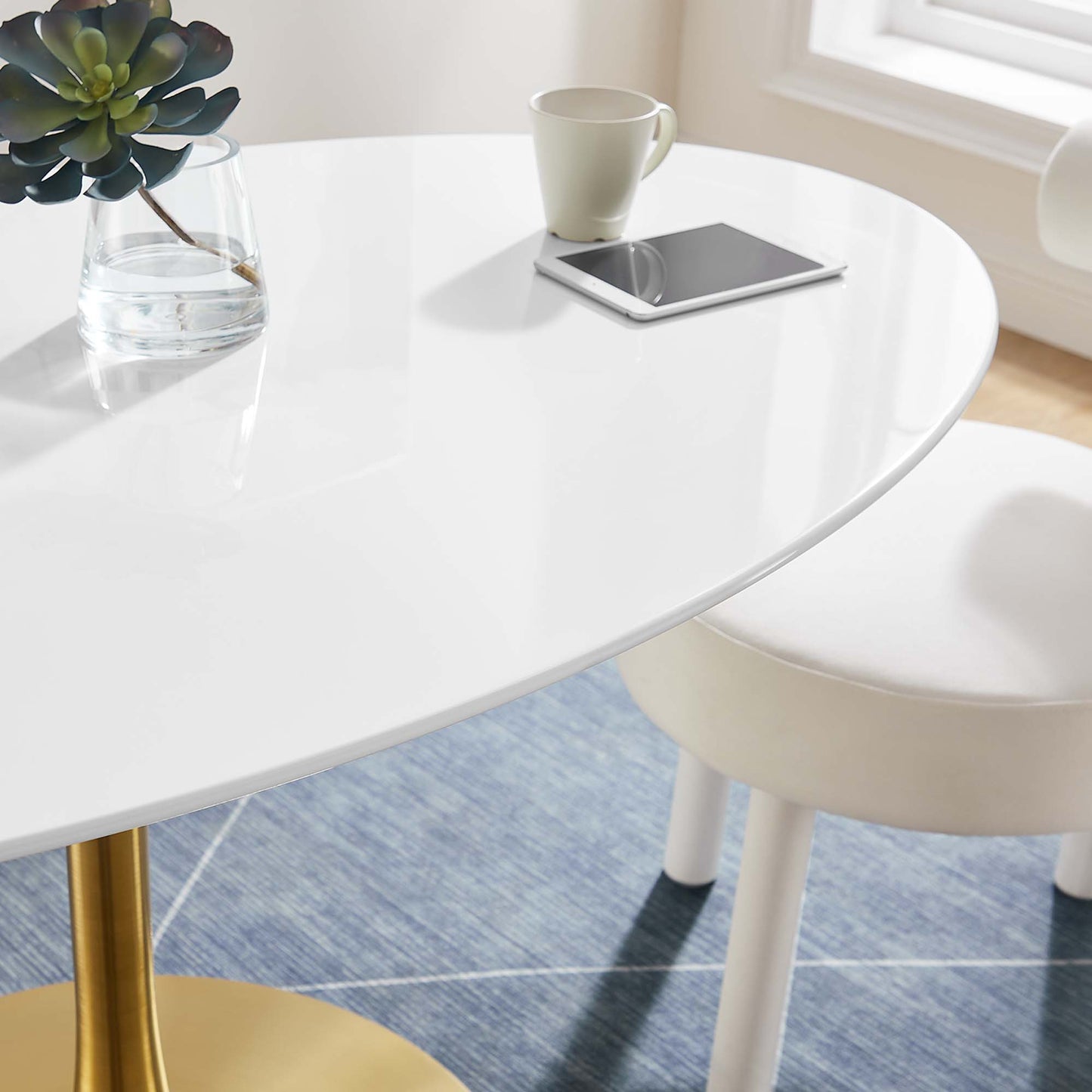 White oval wood dining table with a plant in a glass vase, a white mug and a tablet on top, on a blue rug with a white chair nearby.