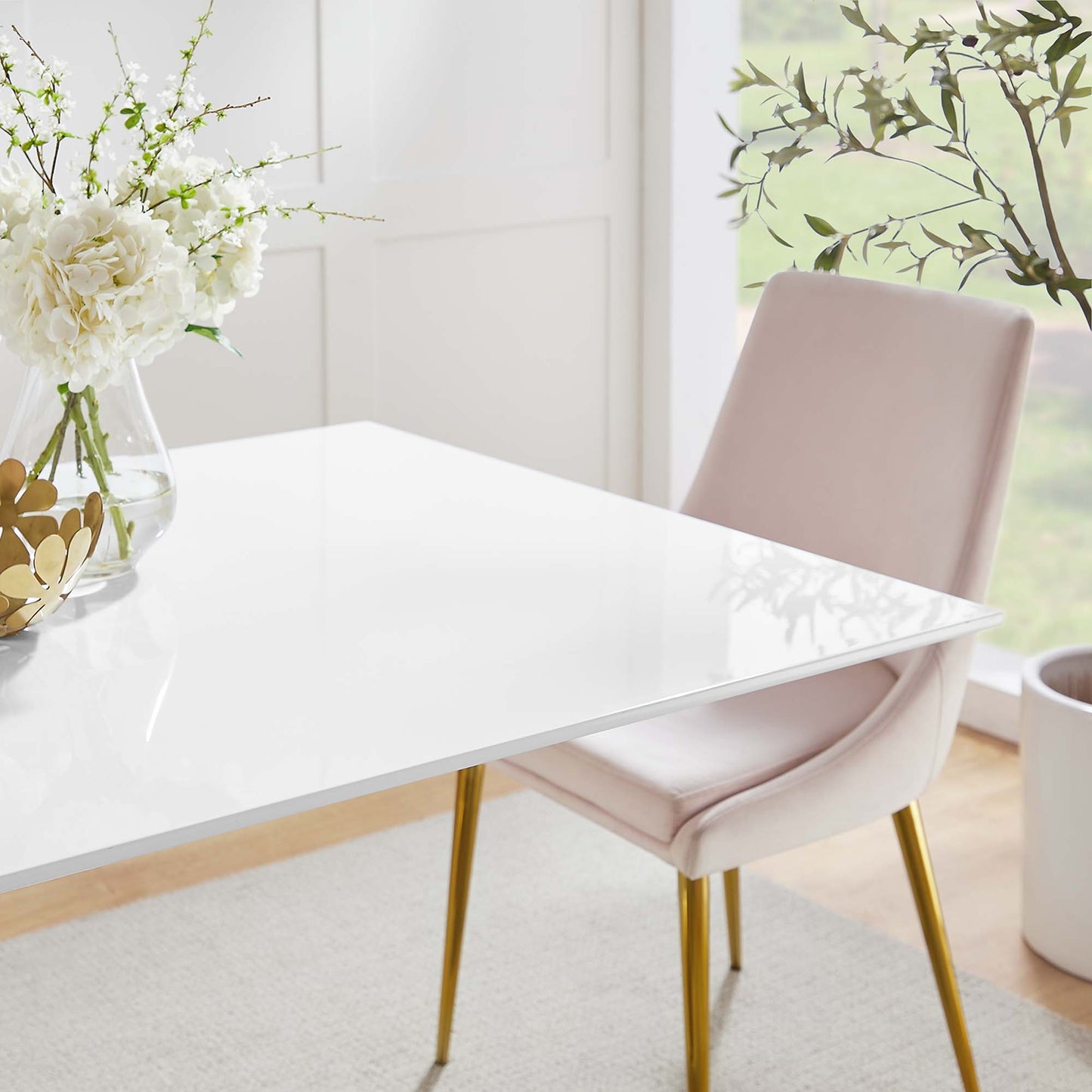 Glossy white table with gold legs, blush pink upholstered chair, vase of white flowers in bright airy dining area