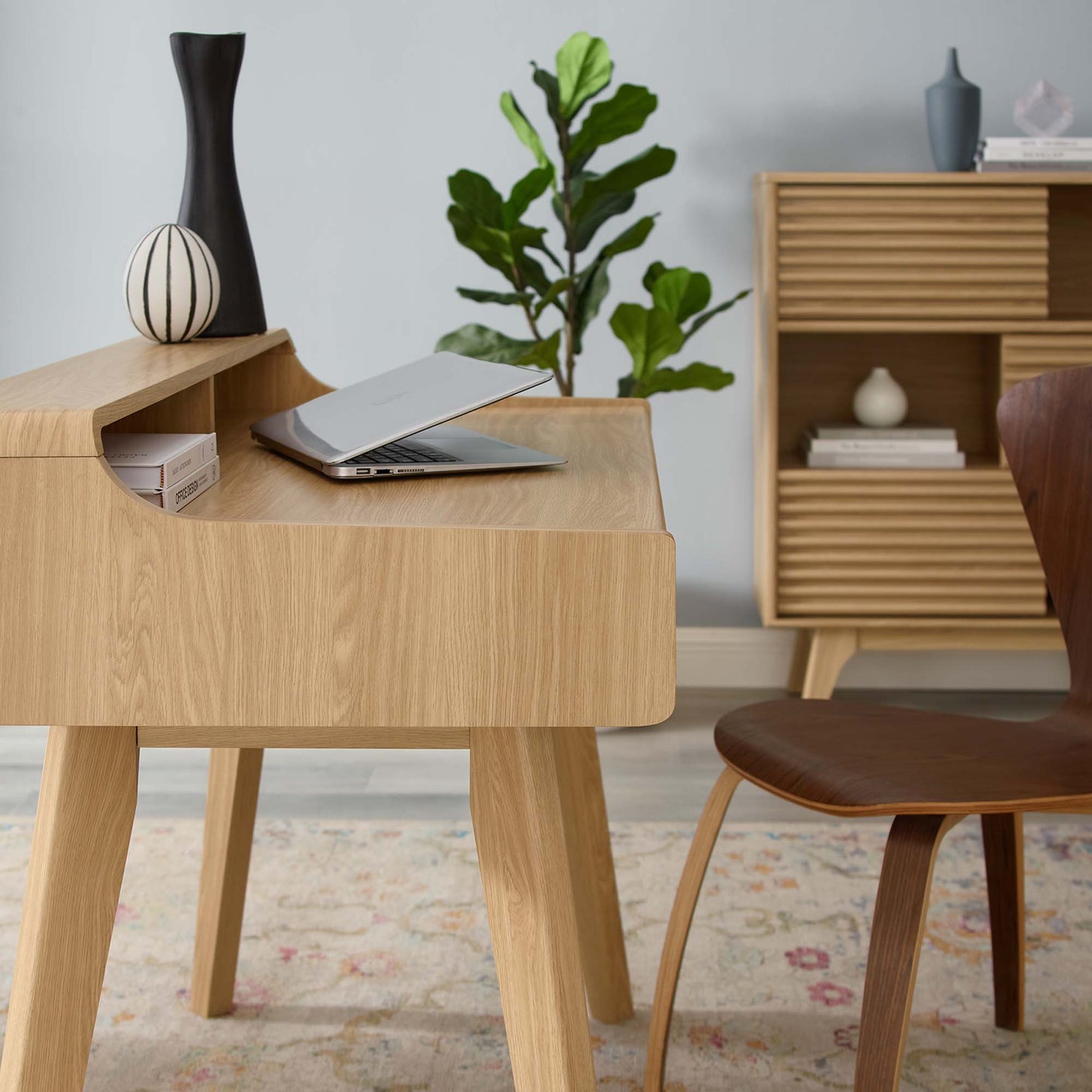 Oak wood desk with rounded edges and slender legs; laptop, black vase, striped ceramic ornament, and a green plant.