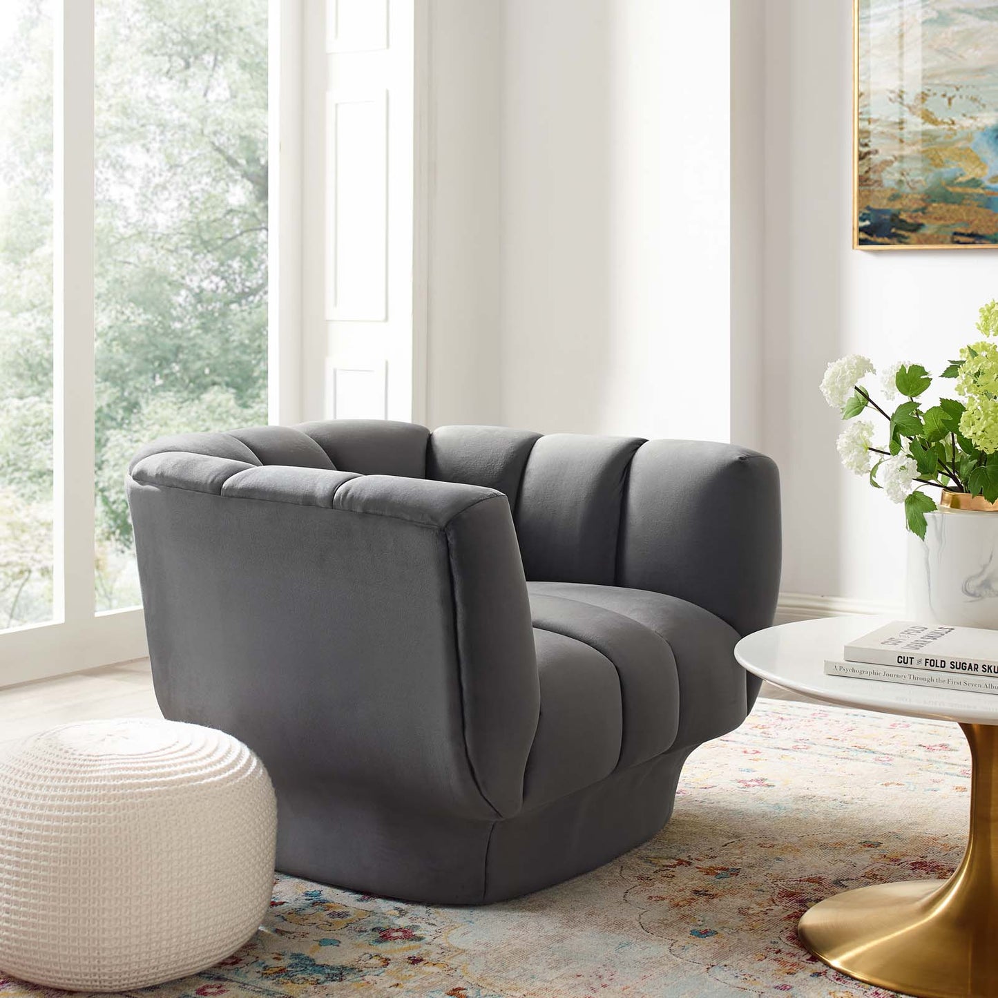 Gray vertical channel tufted velvet armchair in a modern living room with a white pouf, round white table with a gold base.