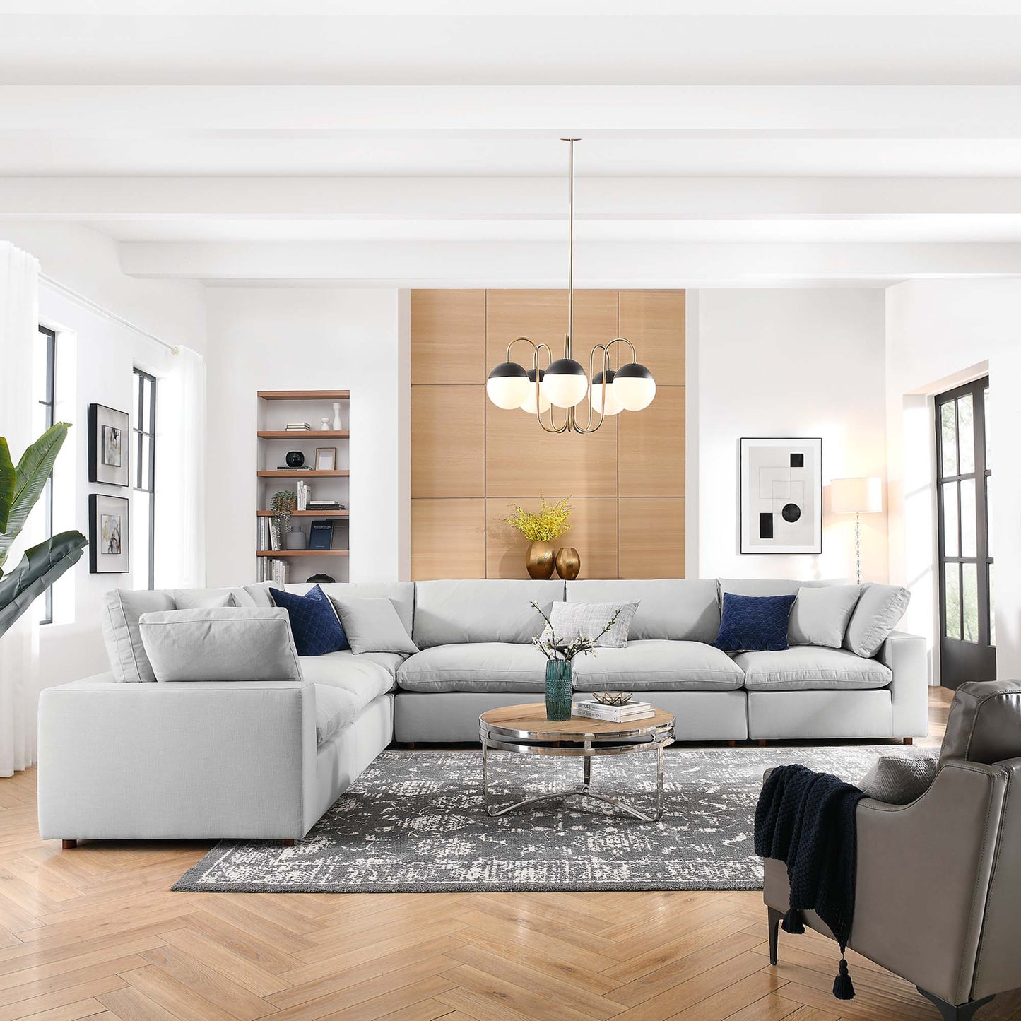 Bright contemporary living room with light gray sectional, round glass table, chandelier, patterned rug and wooden wall unit.