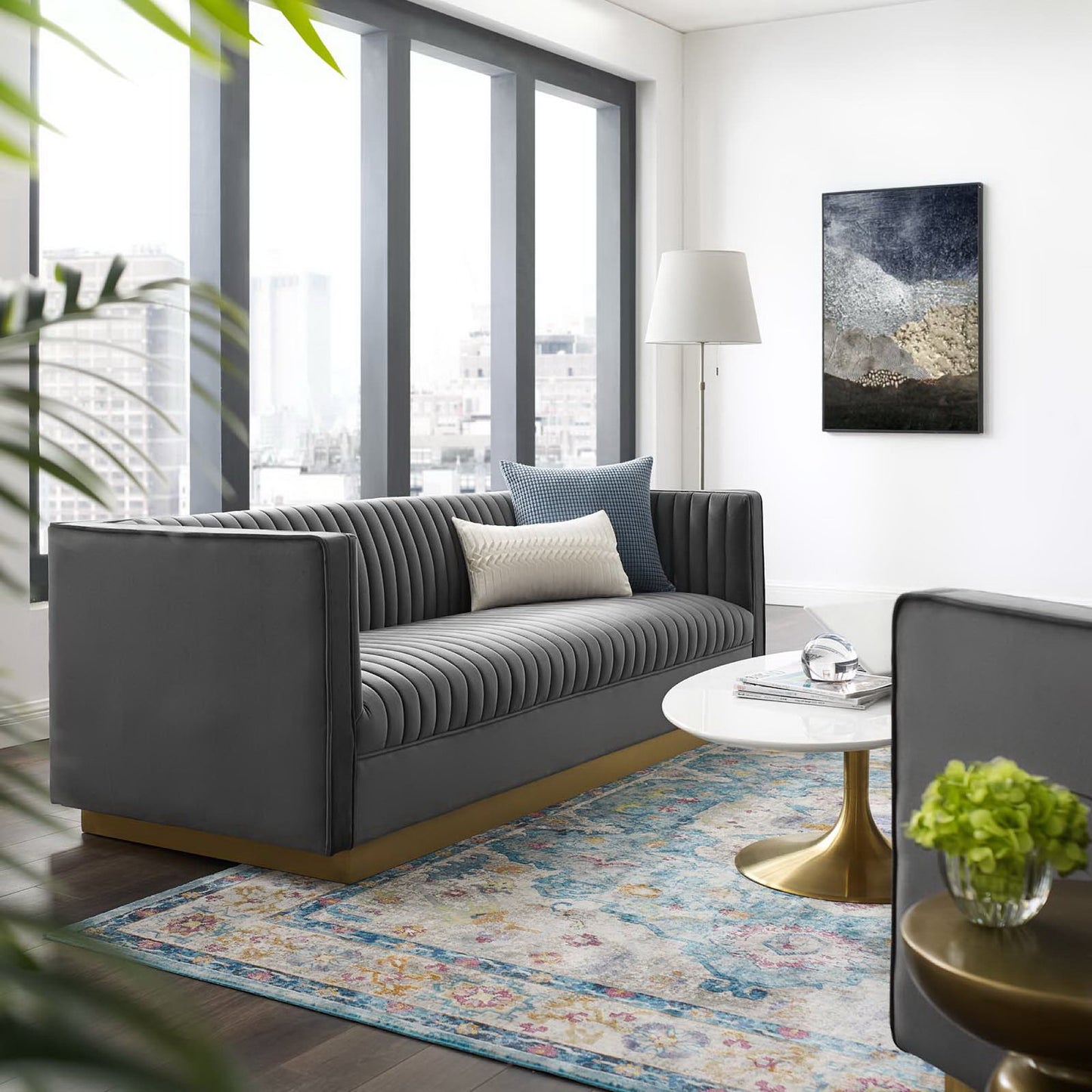 Dark gray vertical-channel tufted velvet sofa with blue/beige pillows in a bright modern living room, round white coffee table with gold base, rug.